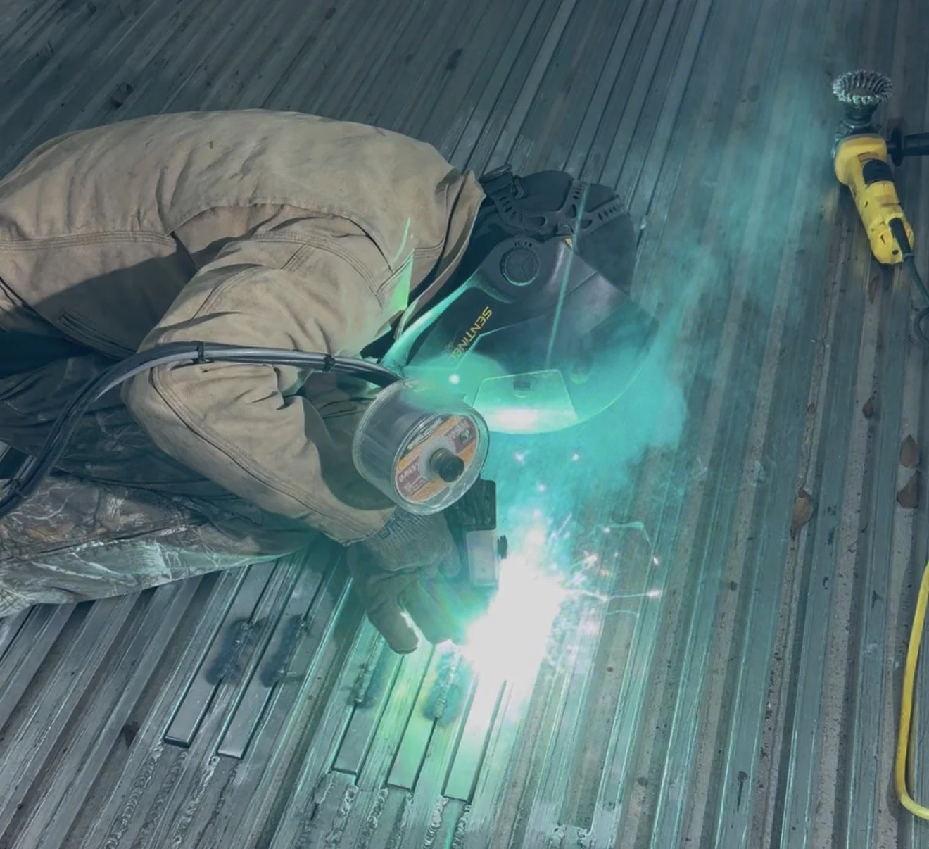 Worker welding metal on a wooden floor, wearing a welding helmet and protective gear, with a yellow power tool nearby.