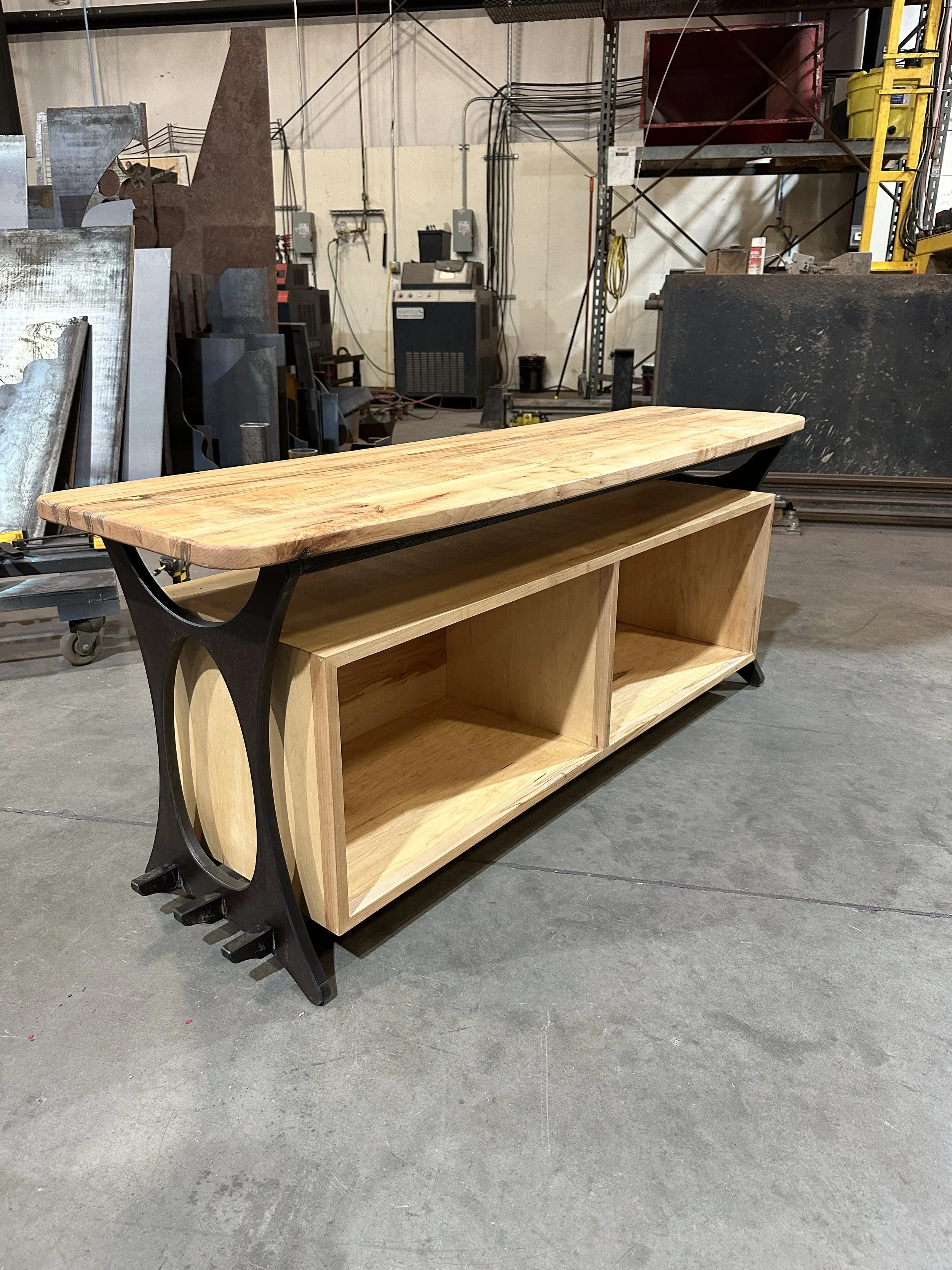 A wooden table with metal legs and a bottom shelf in a workshop setting, with various industrial equipment and metal sheets in the background.