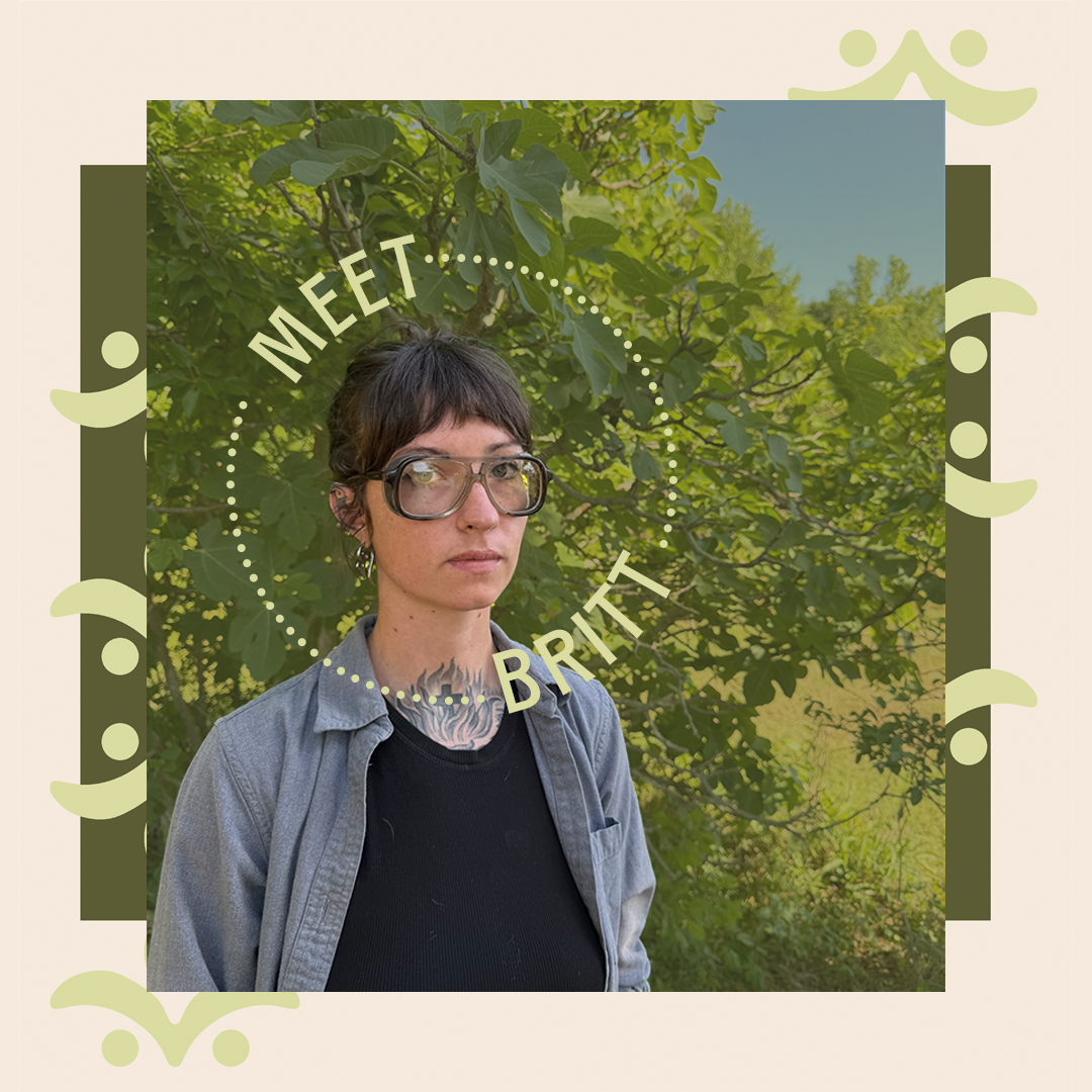 A woman with short dark hair, wearing large glasses, a black t-shirt, and a gray unbuttoned shirt, standing outdoors in front of lush green foliage.