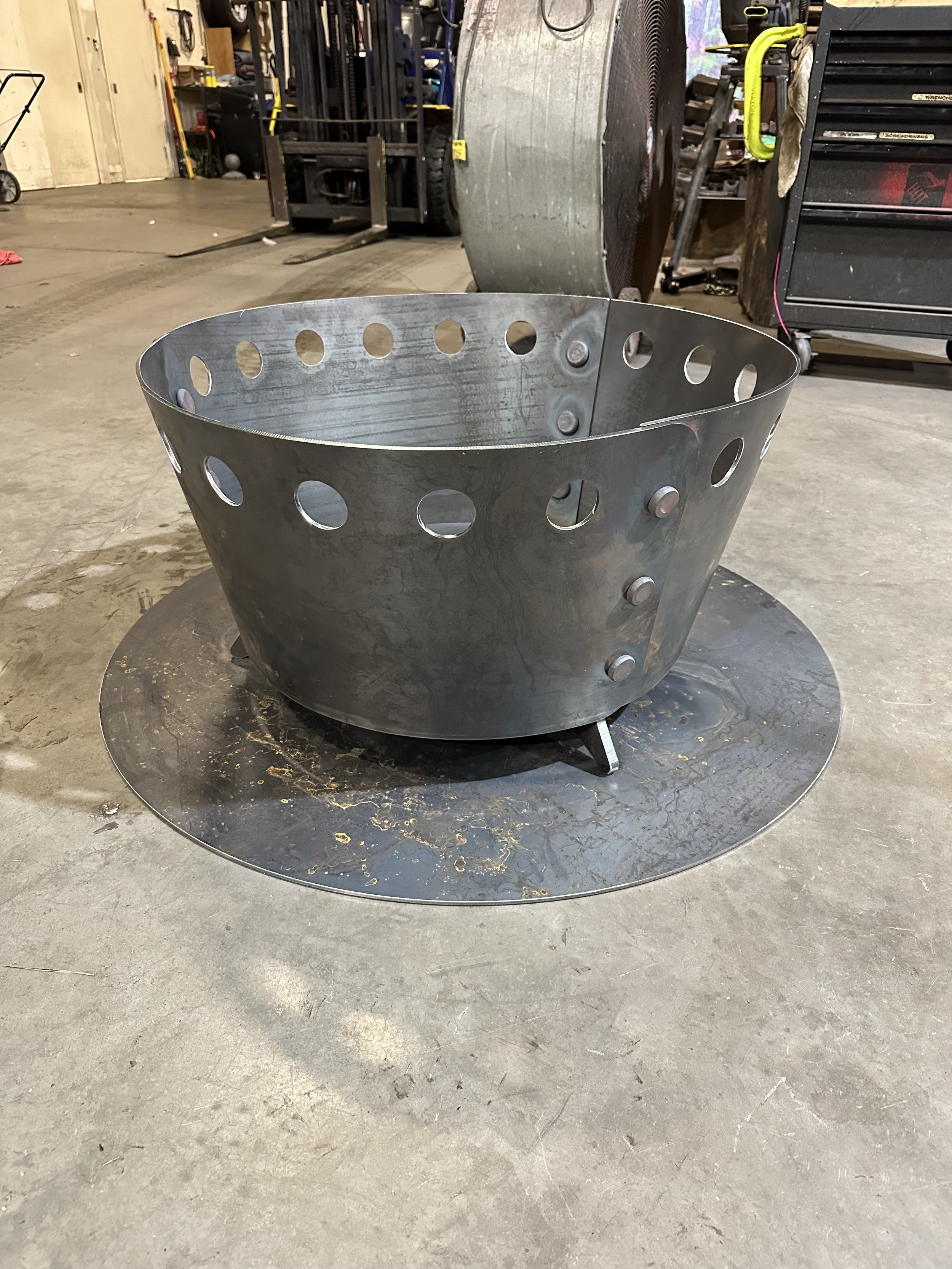 Metal fire pit with circular holes, sitting on a concrete garage floor, with a background of tools and equipment.