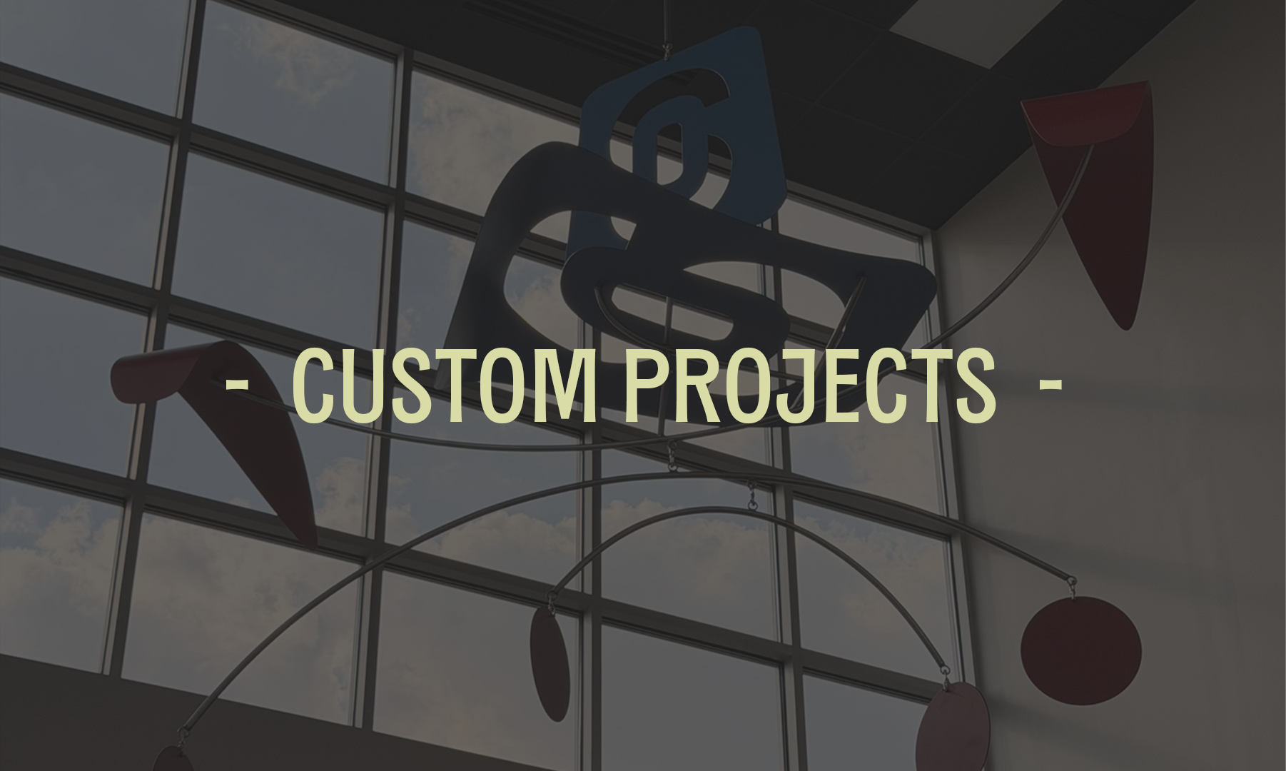 Interior view of a modern building with large window panels. Hanging from the ceiling are abstract art sculptures with curved shapes and circular elements. Text overlay reads '- Custom Projects -'.