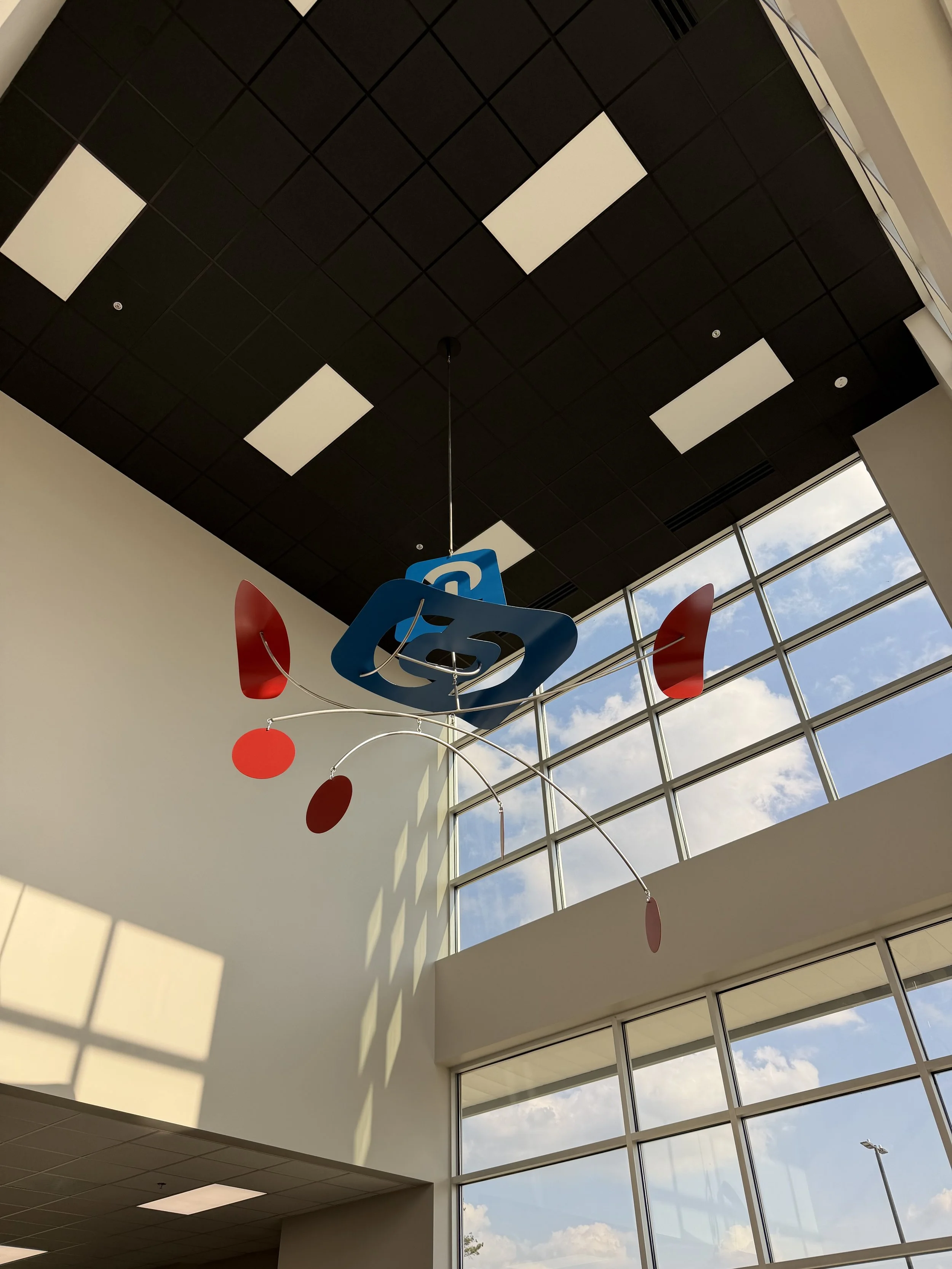 Interior view of a building with a black grid ceiling, large windows showing a partly cloudy sky, and a colorful electronic display sculpture hanging from the ceiling.