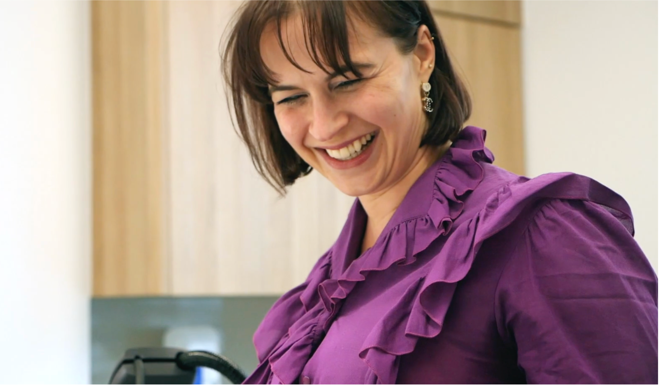 A woman with short brown hair smiling, wearing a purple ruffled blouse and pearl earring, in an indoor setting.