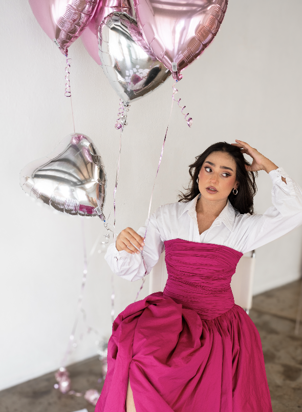 A young woman with dark hair and hoop earrings, wearing a white blouse and a bright pink dress, standing in front of a white wall. She is holding silver and pink heart-shaped balloons and has one hand on her head, looking off to the side.
