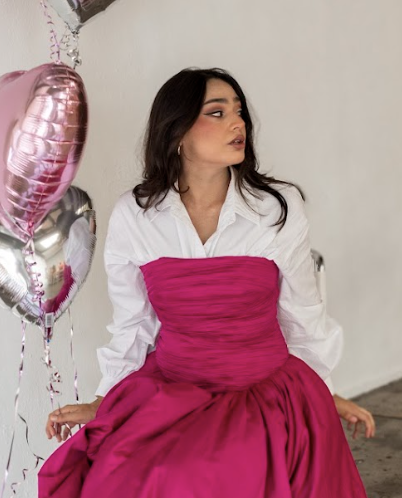 A woman wearing a white shirt with a pink fabric overlay, sitting next to pink and silver heart-shaped balloons.