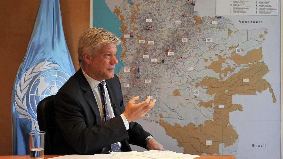 A man in a suit gesturing while sitting at a table in front of a large map of Colombia and surrounding countries, with a United Nations flag in the background.
