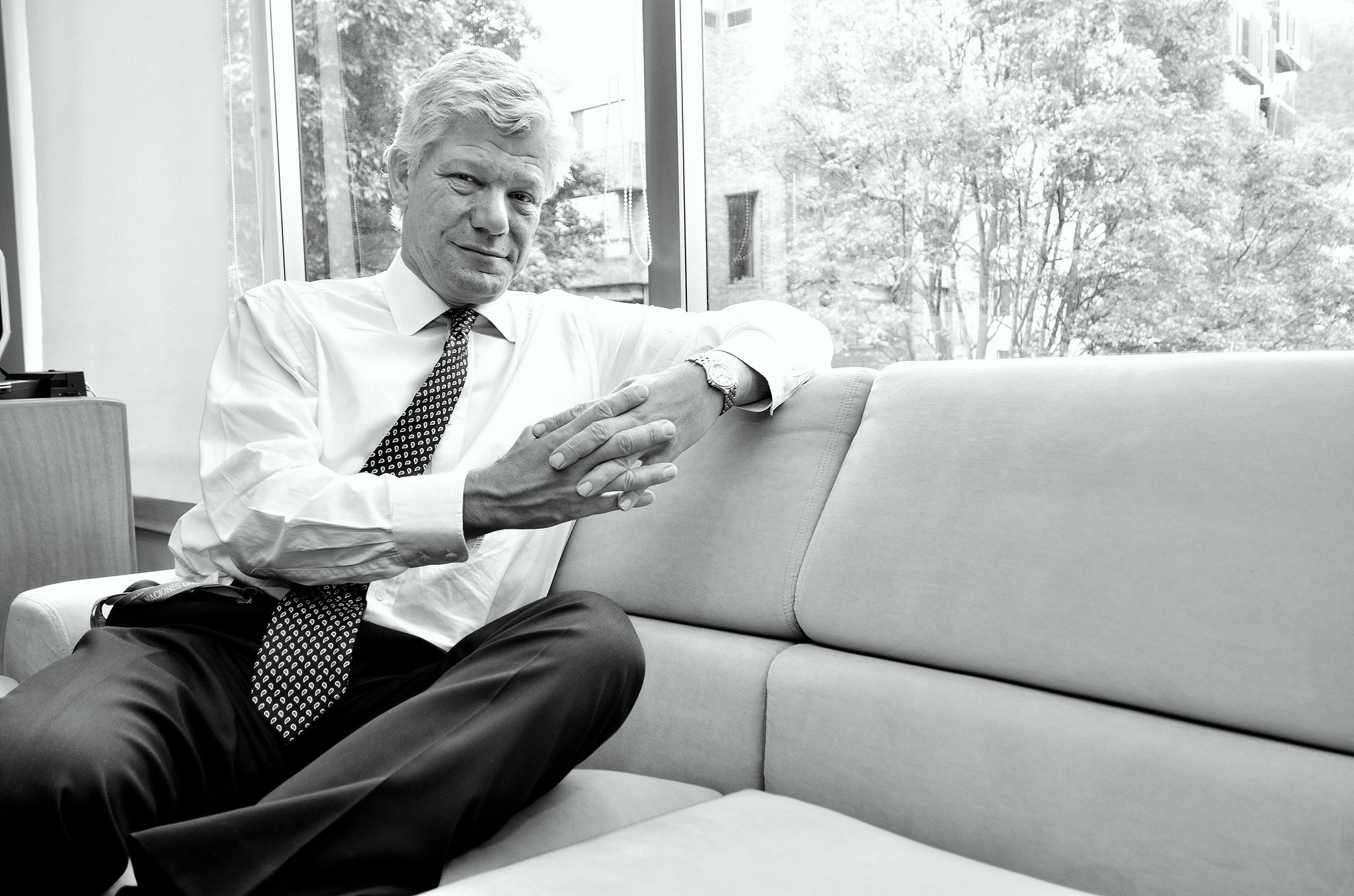 A black and white photo of a man with white hair in a white shirt and patterned tie sitting on a couch, looking at the camera with his hands clasped, in an office or waiting room with large windows and trees outside.