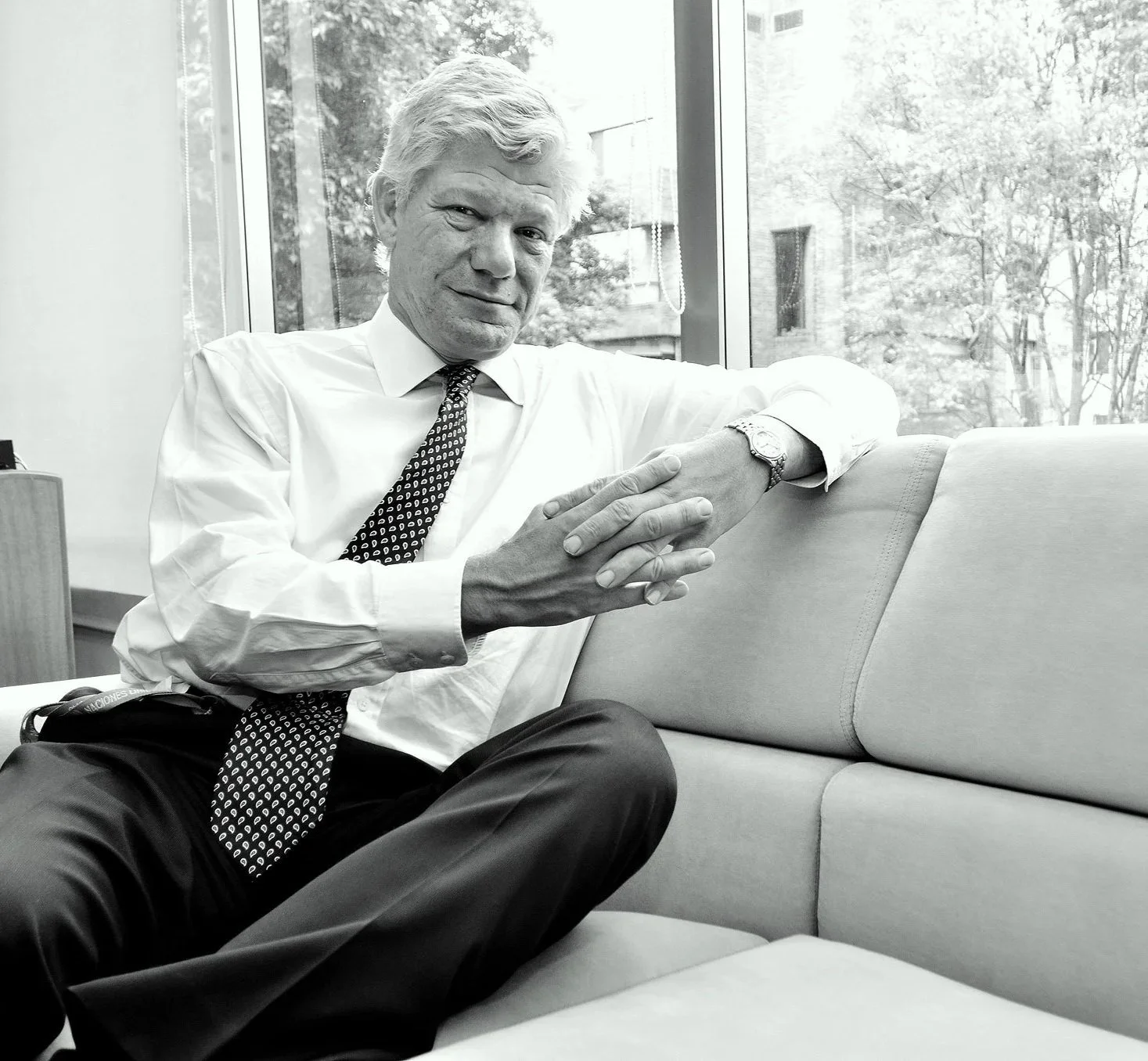 A black and white photo of a man sitting on a couch indoors with a large window behind him. He has light-colored hair, is wearing a white shirt, patterned tie, and a watch. He is leaning slightly forward with his hands clasped and smiling gently at the camera.