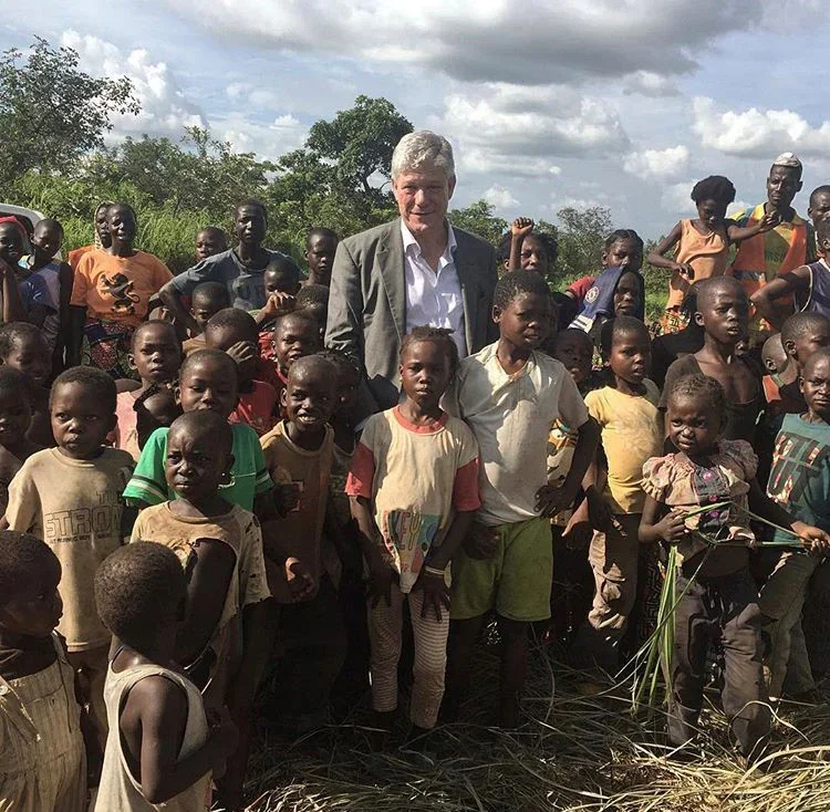 A man with gray hair and a blazer surrounded by several children outdoors in a rural area with trees and grass, under a partly cloudy sky.