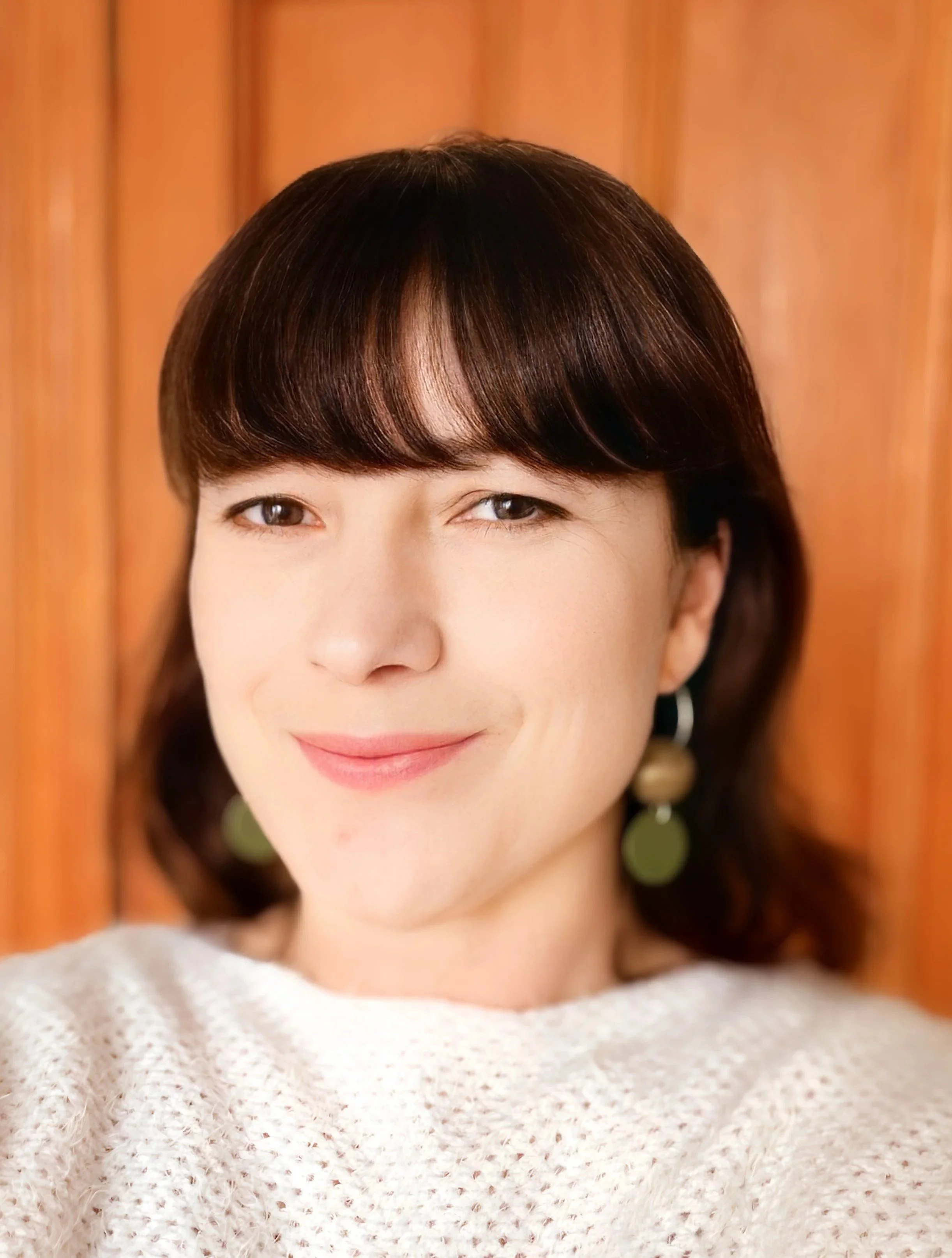 A woman with short, dark brown hair and light makeup, smiling softly, wearing a cream-colored textured sweater and large earrings, standing in front of a wooden panel background.