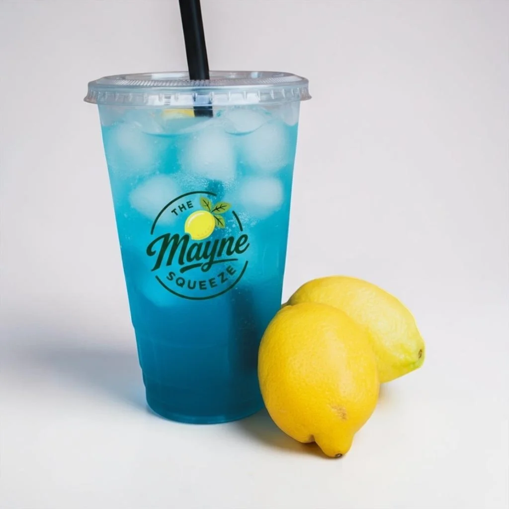 A blue lemonade in a clear plastic cup with ice and a black straw, placed next to two fresh lemons, on a plain white background.