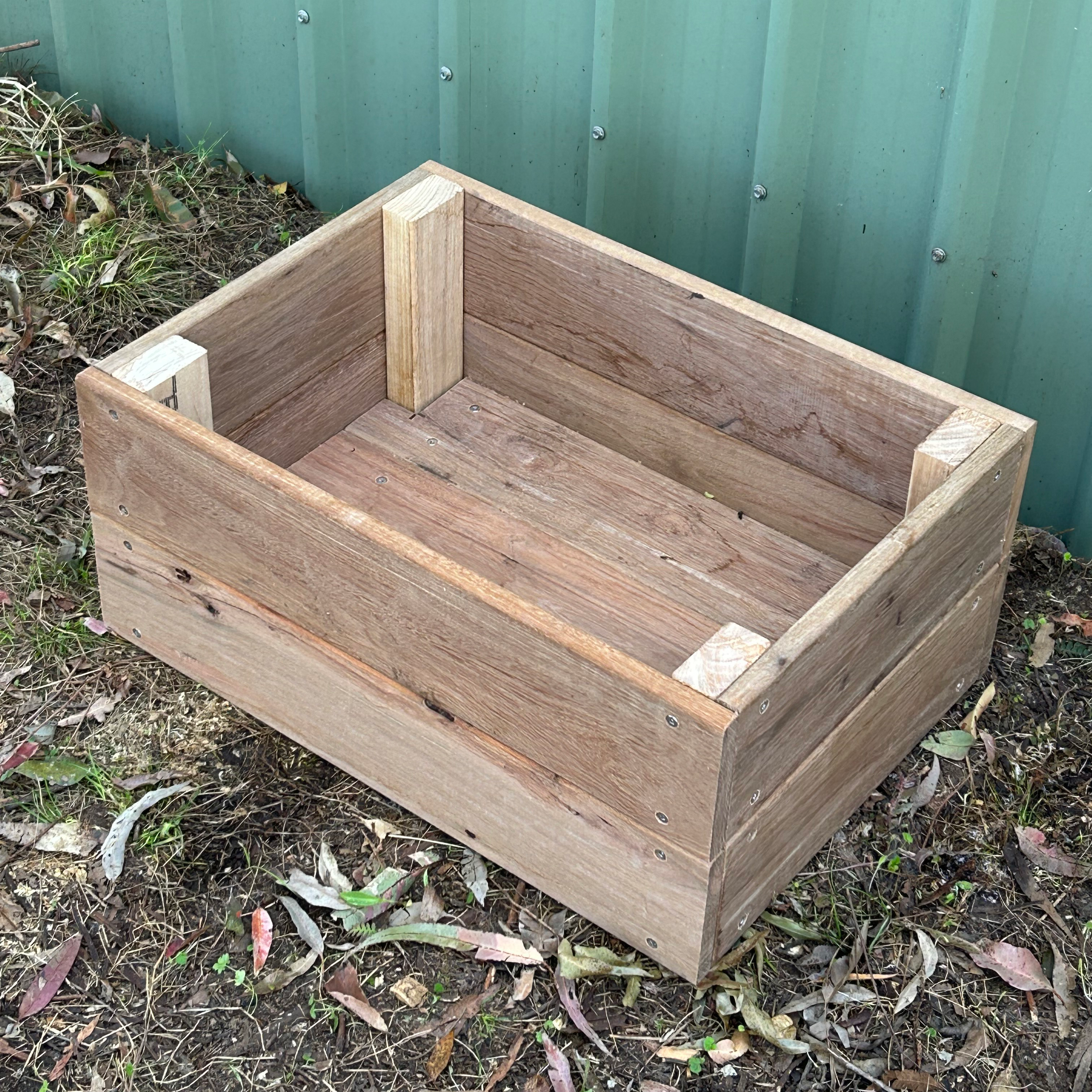 A wooden planter box with soil and plants on the ground next to a green metal fence.