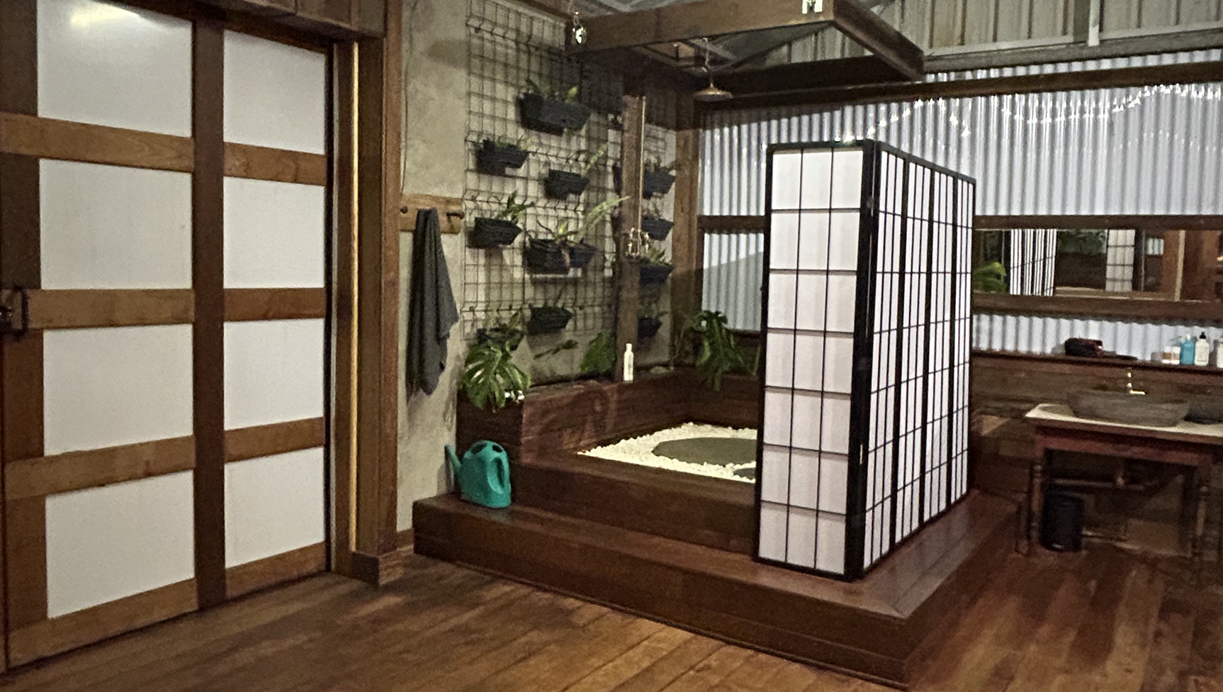 Indoor space with wooden flooring, a raised wooden platform containing a small gravel garden with a black circular feature, potted plants, and a folding screen partition. Behind, a wire grid wall displays hanging plant pots and a cloth towel, with a corner shelf holding various items.