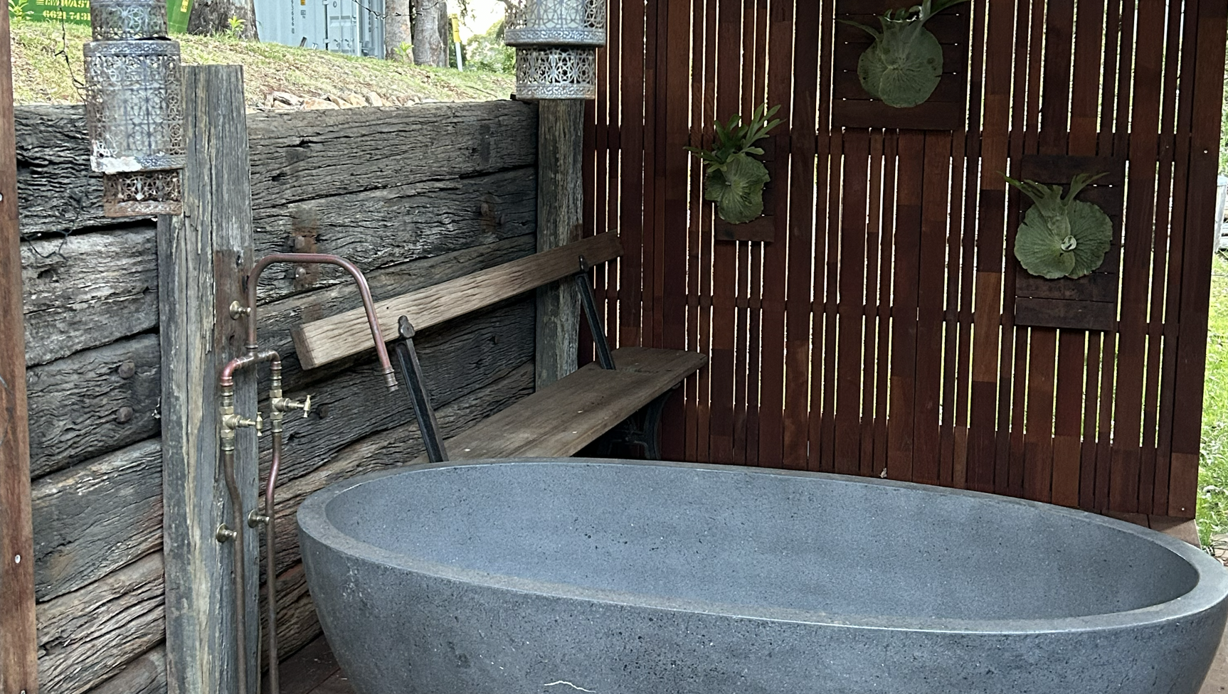 A rustic outdoor bath area with a large stone bathtub, a wooden bench, a wooden privacy fence with mounted green leaves, and decorative metal lanterns.
