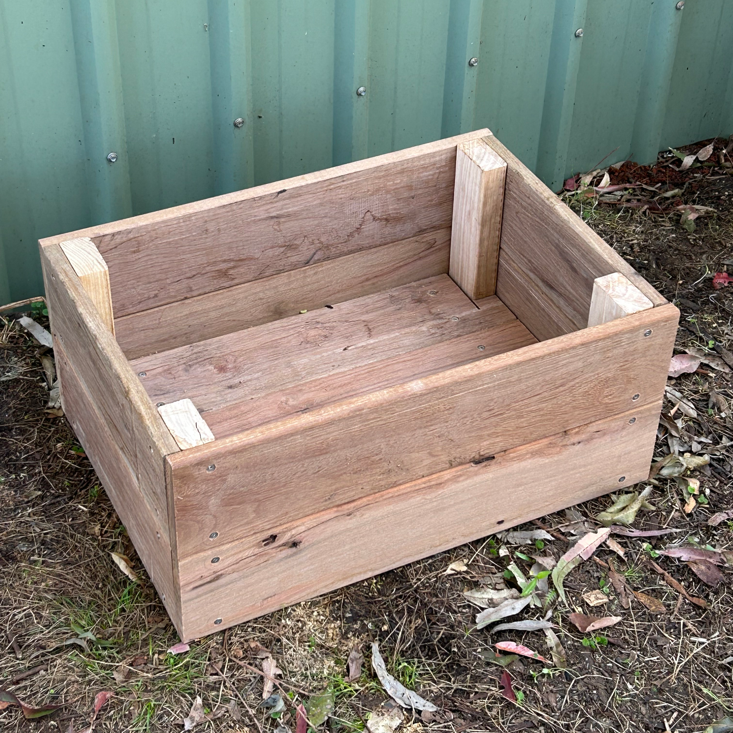 A wooden garden bed frame outdoors next to a green metal wall, with soil and fallen leaves on the ground.