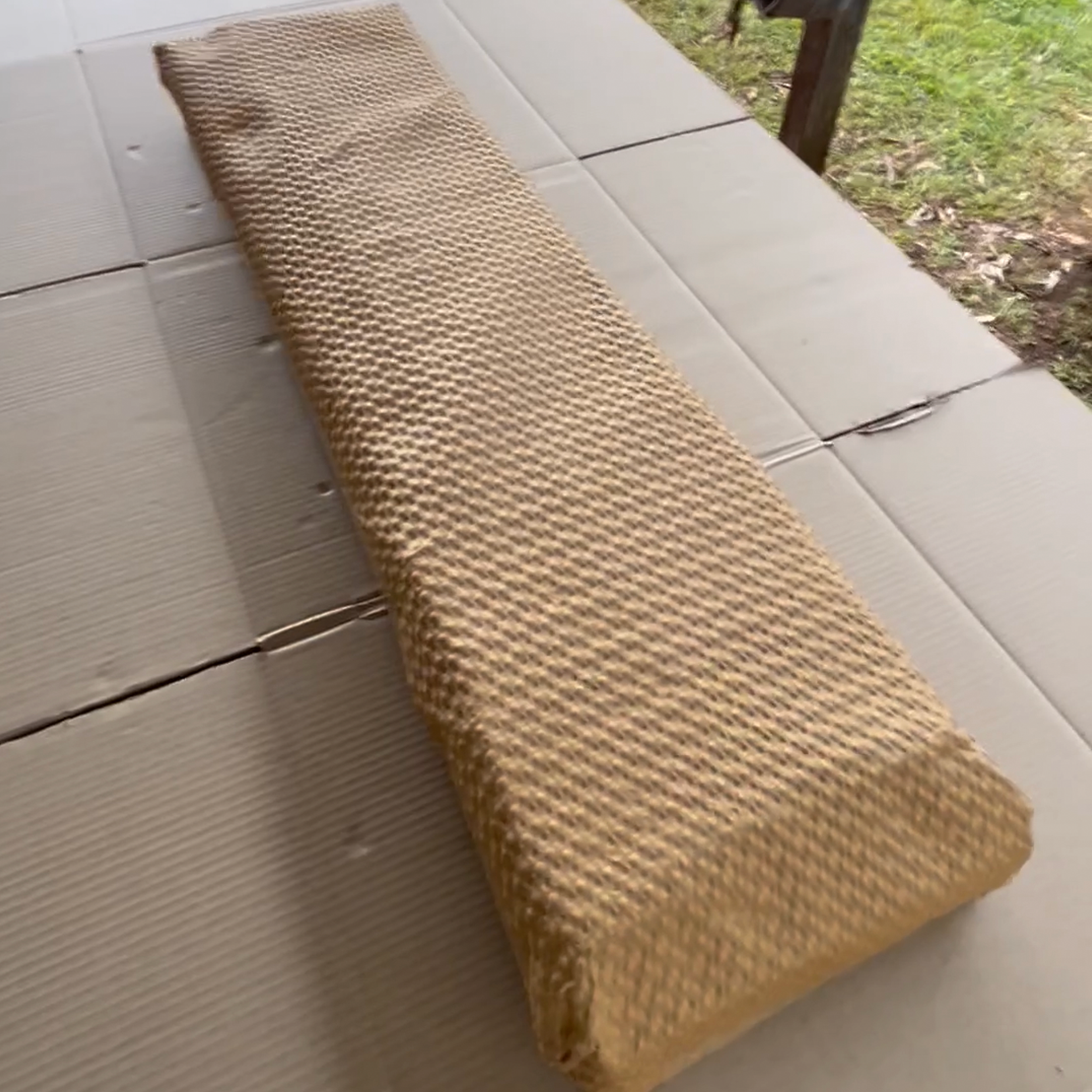 A close-up of a light brown, textured mat on a white outdoor tiled surface, with grass and a wooden structure visible in the background.