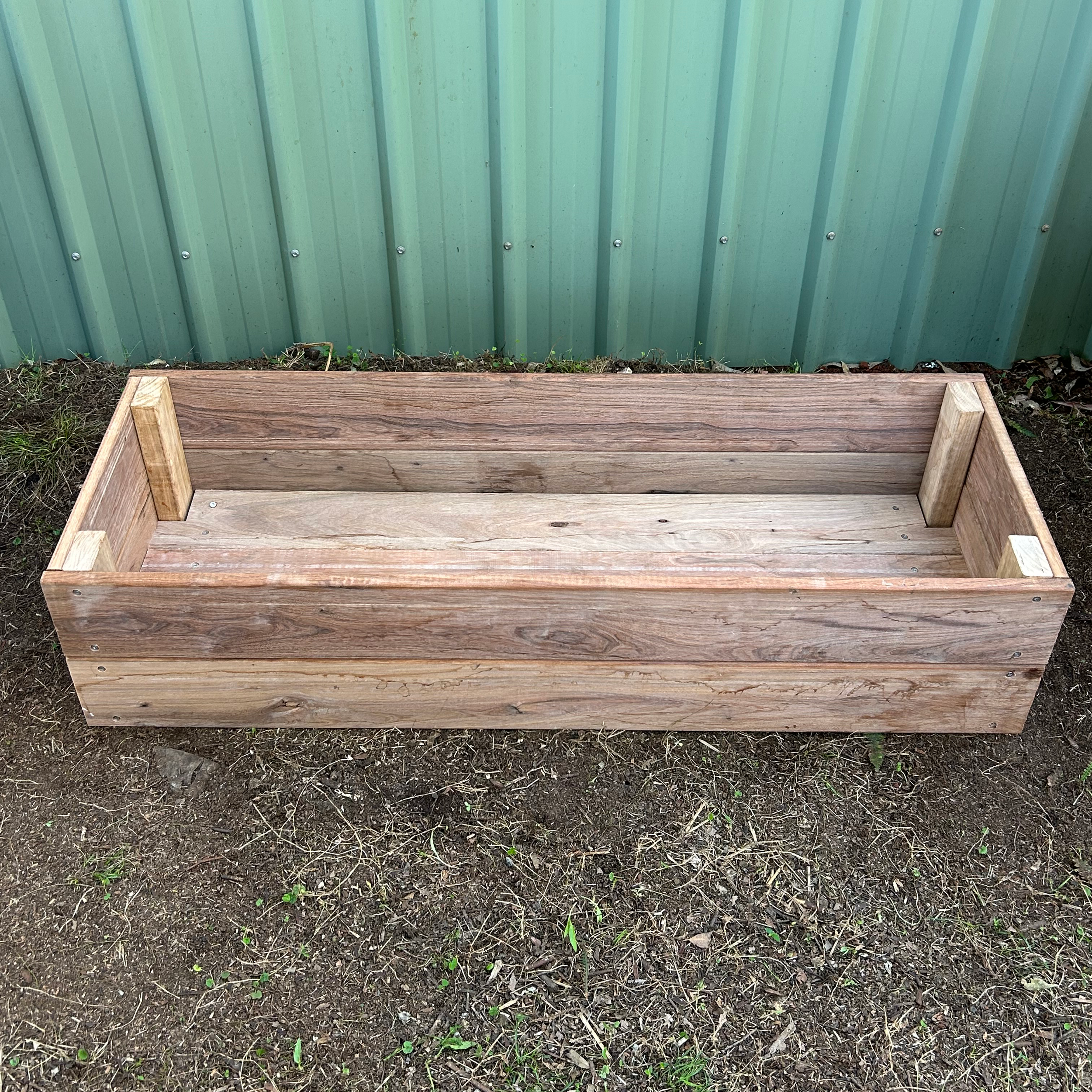 A wooden garden bed frame positioned on dirt ground next to a green metal fence.