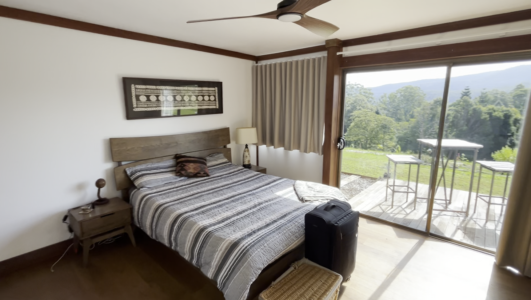 Bedroom with wooden bed and striped bedding, nightstand with lamp and phone, black suitcase, framed artwork above headboard, sliding glass door to balcony with outdoor table and chairs, view of green trees and mountains outside.