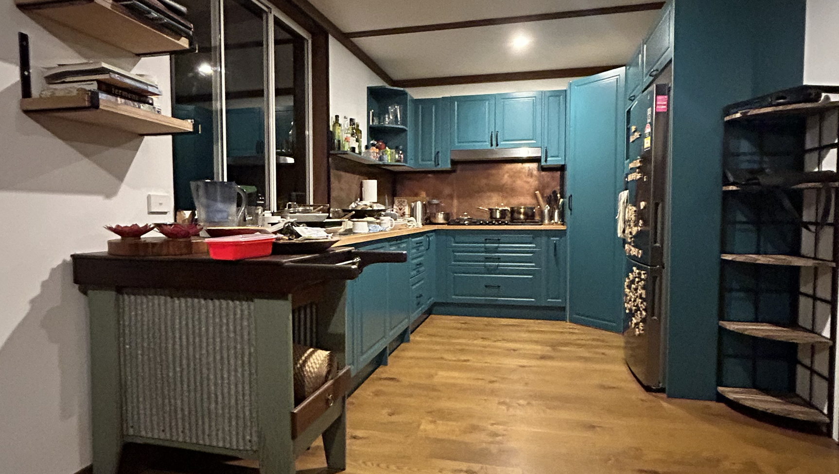 A cluttered kitchen with teal cabinets, wooden countertops, and a wooden floor. The kitchen has a variety of dishes, containers, and cooking utensils on the counters. There is a small cart in the foreground with bowls and a woven basket, and open shelves on the left holding books. Black metal shelves on the right hold items. A stainless steel refrigerator is visible on the right, and above the countertops are glass-fronted cabinets and a brown backsplash.