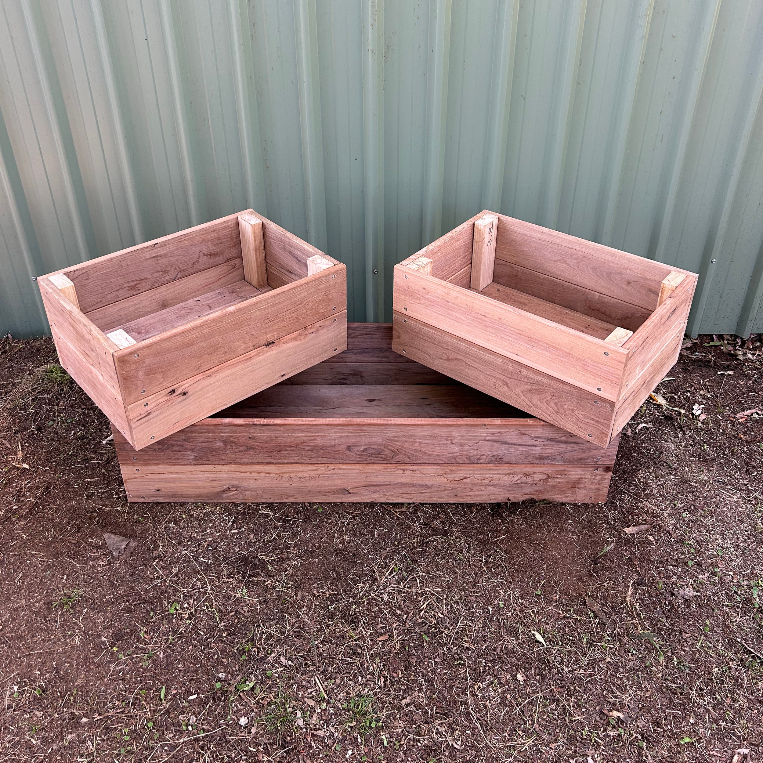 Two unfinished wooden planters on a patch of dirt soil, positioned in front of a green metal wall.