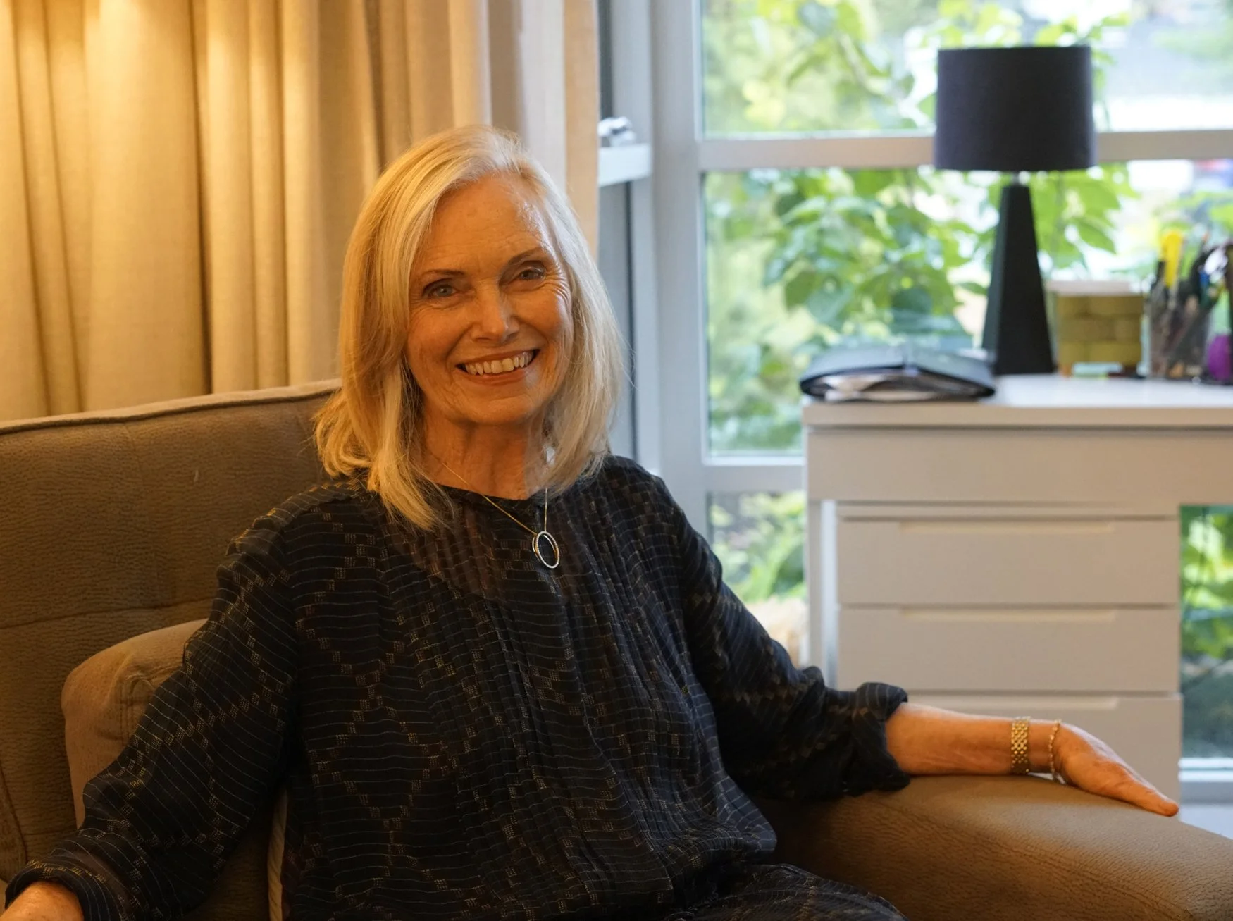 A middle-aged woman with blonde hair smiling while sitting on a beige armchair near a window with greenery outside. She is wearing a black patterned top and jewelry, including a necklace and a bracelet, in a brightly lit room with beige curtains and a white drawer.