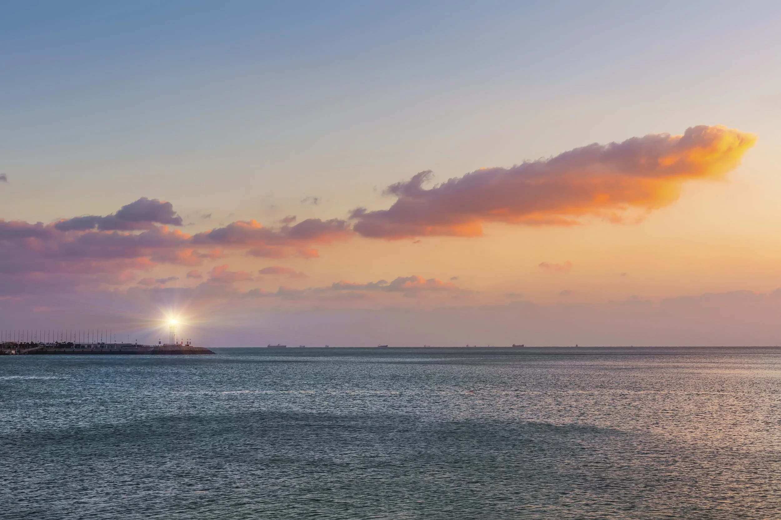 Sunset view over the ocean with a pier and ships in the distance, and colorful clouds in the sky.