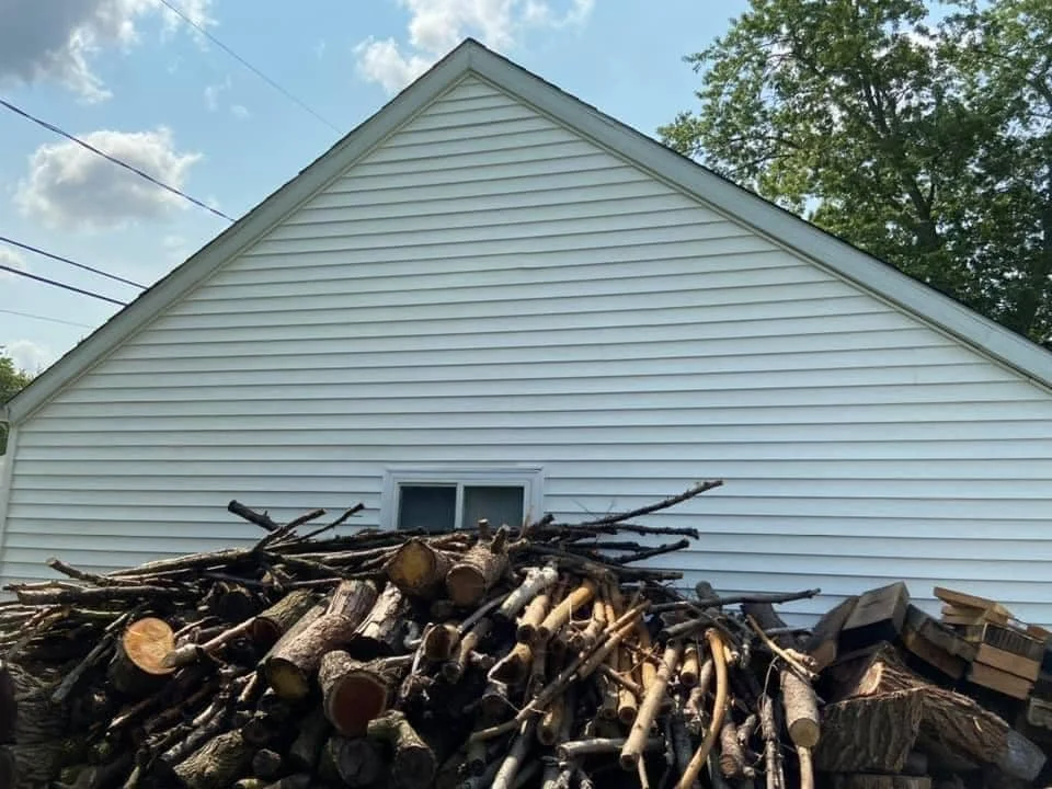 Stack of cut logs and branches in front of a white house with vinyl siding and a small window; blue sky with clouds and green trees in the background.