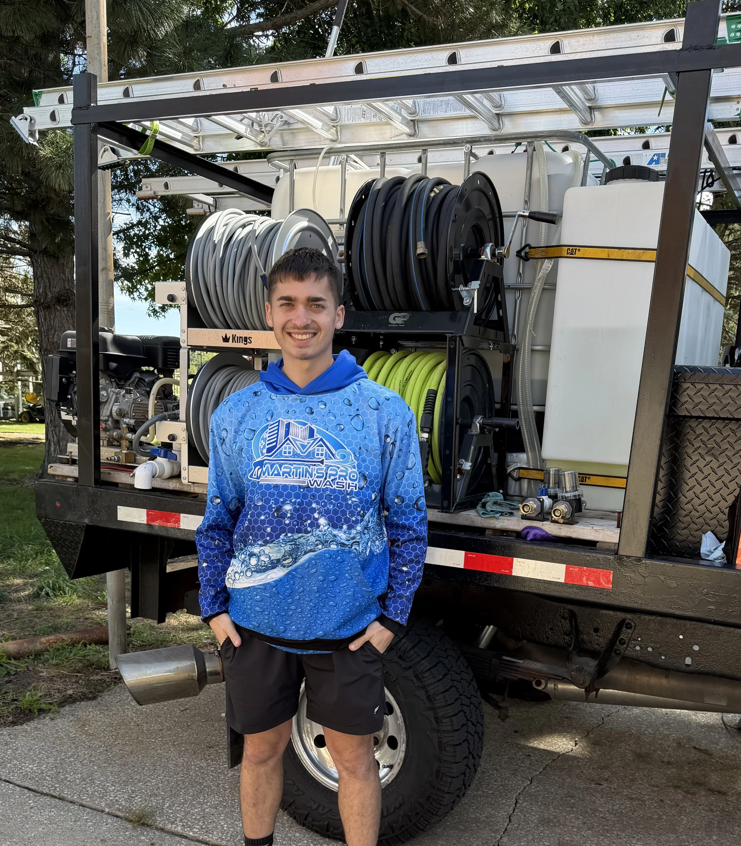 A smiling man stands in front of a utility truck with equipment and hoses, wearing a blue running jacket and black shorts, outdoors with trees in the background.