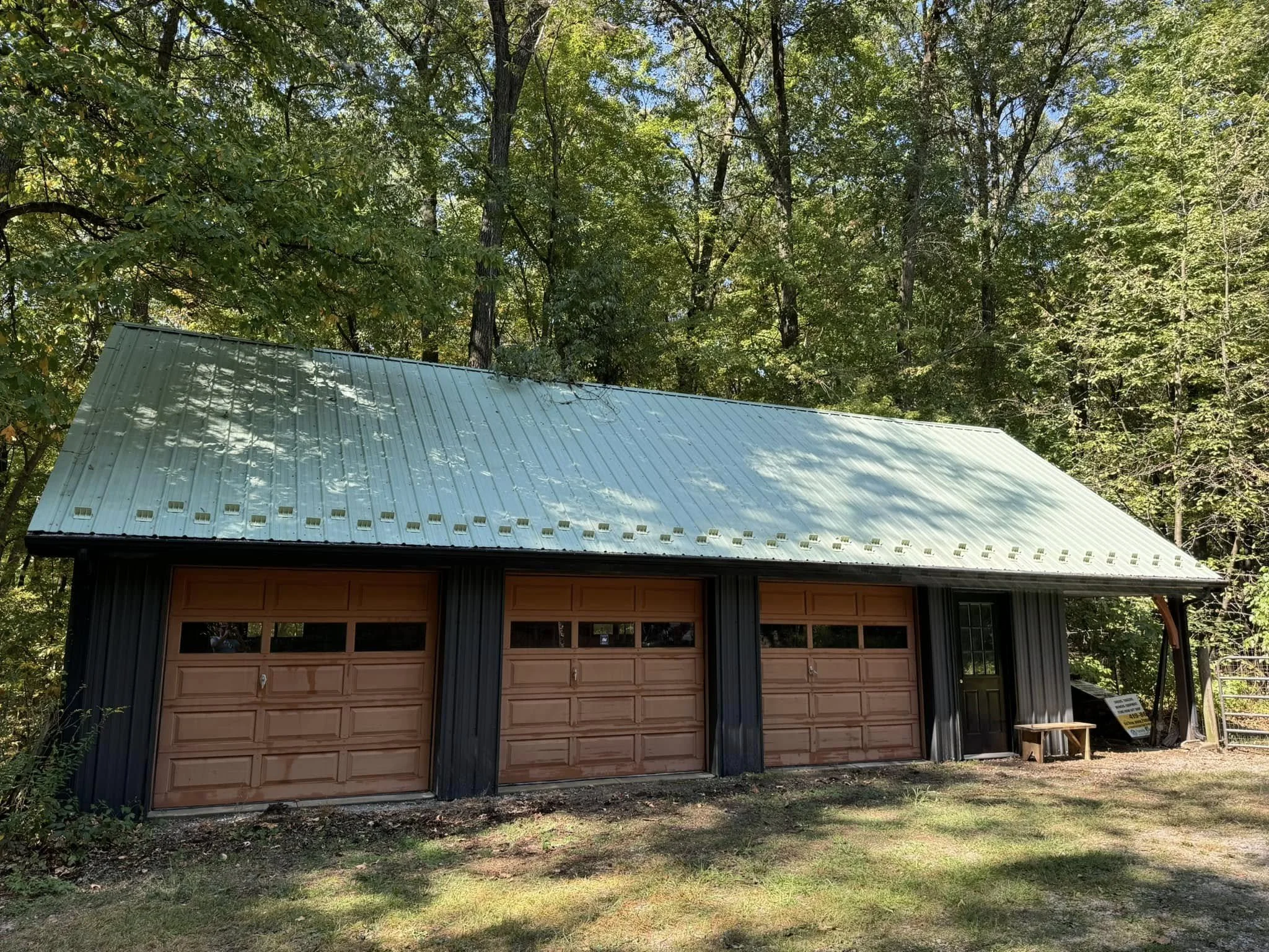 A metal-roofed building with a large garage door, set in a wooded area with tall trees and green foliage surrounding it.