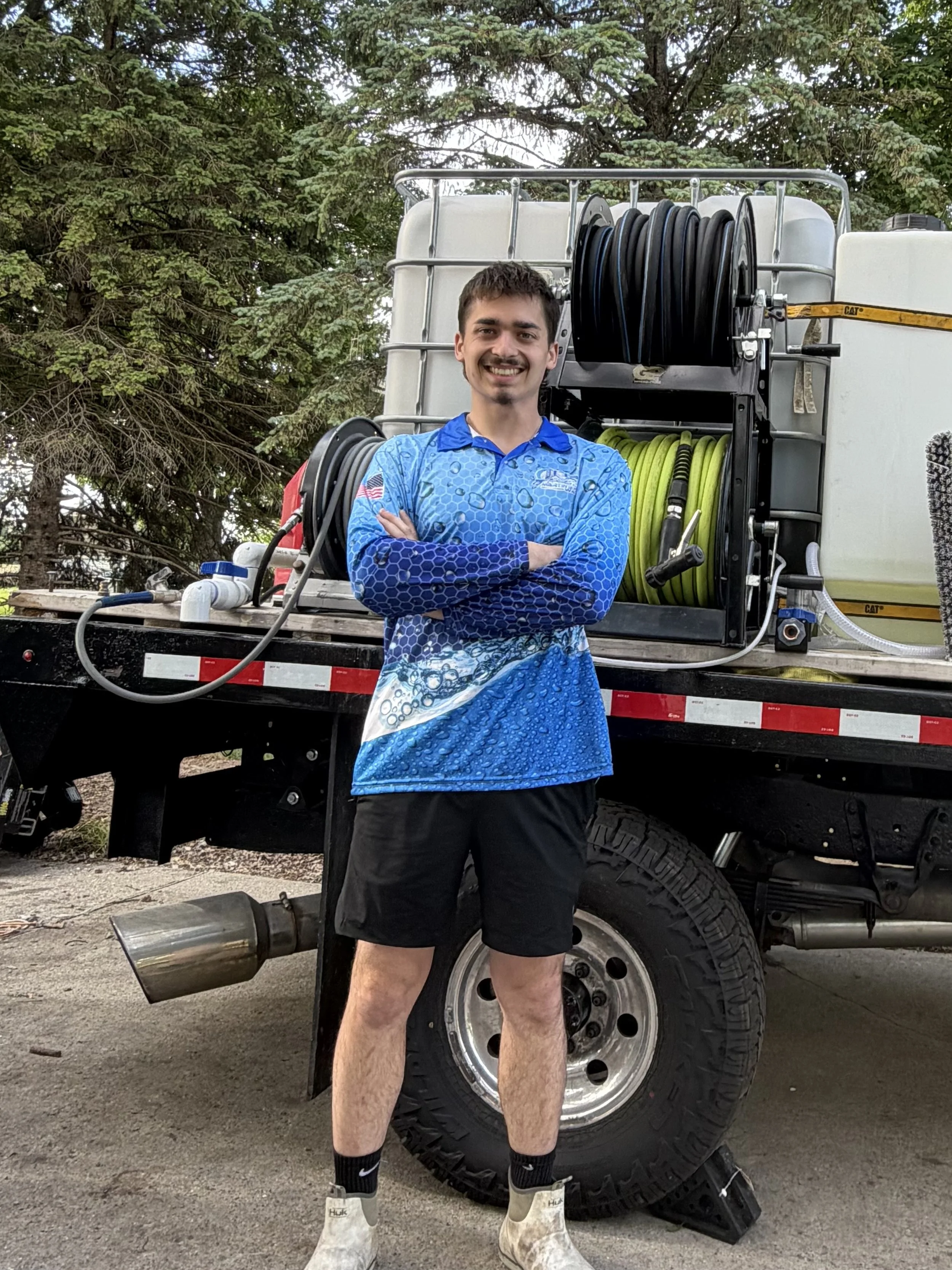 A young man standing in front of a large truck with hoses and equipment, wearing a blue patterned shirt, shorts, and white boots, outdoors with trees in the background.