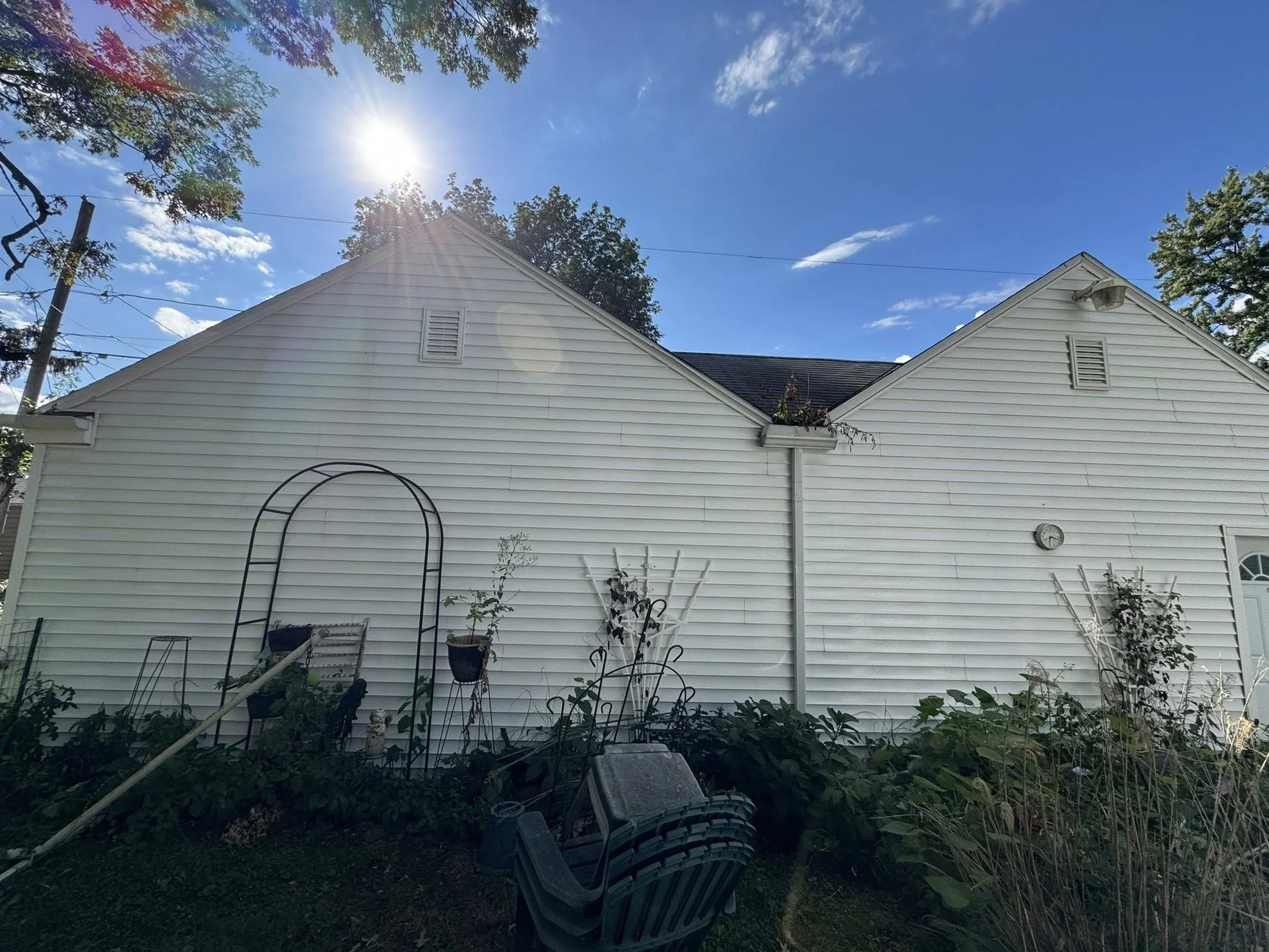 Backyard with white house siding, plants, garden tools, and gardening supplies, under a clear blue sky with the sun shining.