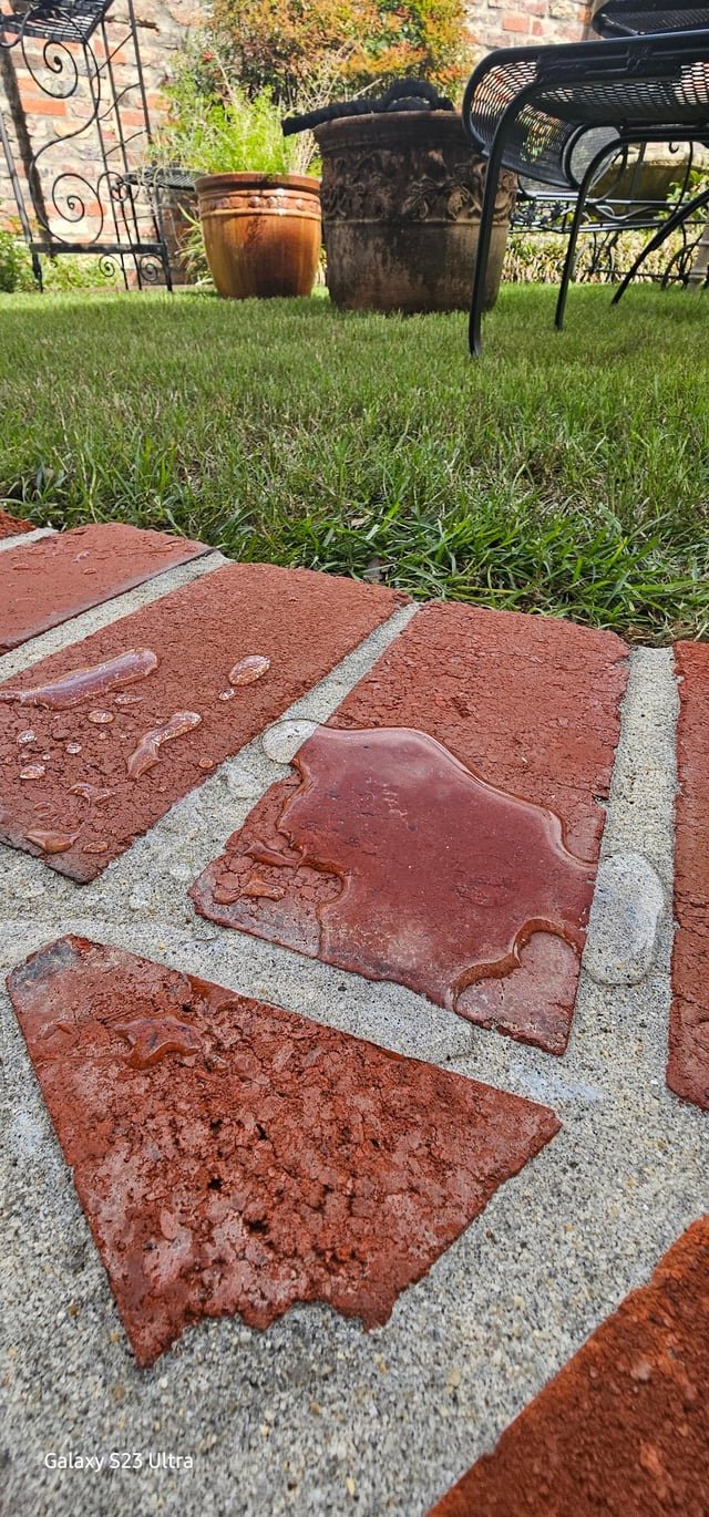 Close-up of wet red brick pavers on a patio with grass in the background and patio furniture. Pavers and grass are near a brick wall with plants and large ceramic pots.