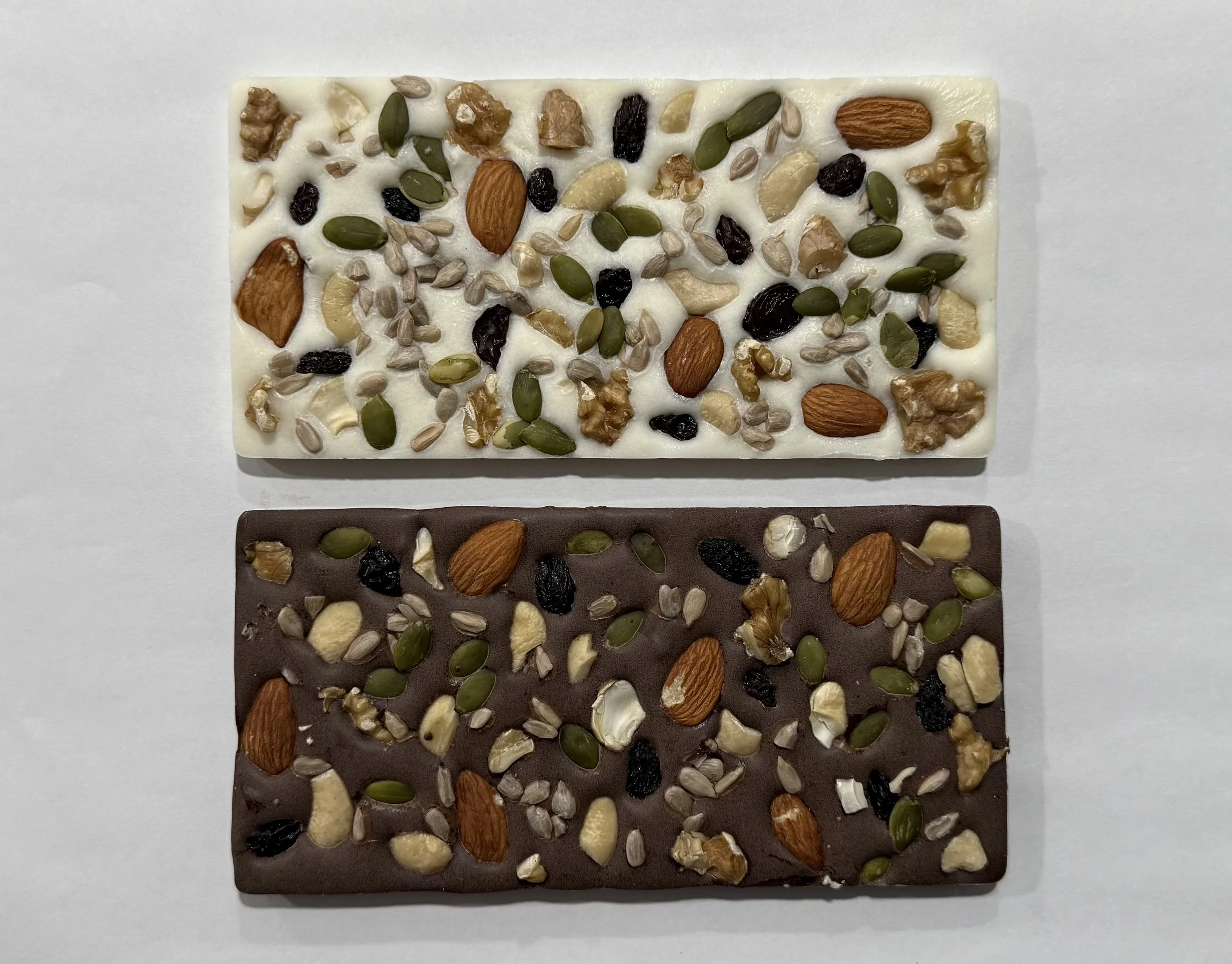 Close-up of a white chocolate bar topped with mixed nuts and dried fruit, including almonds, walnuts, pumpkin seeds, sunflower seeds, and black raisins, on a white background.