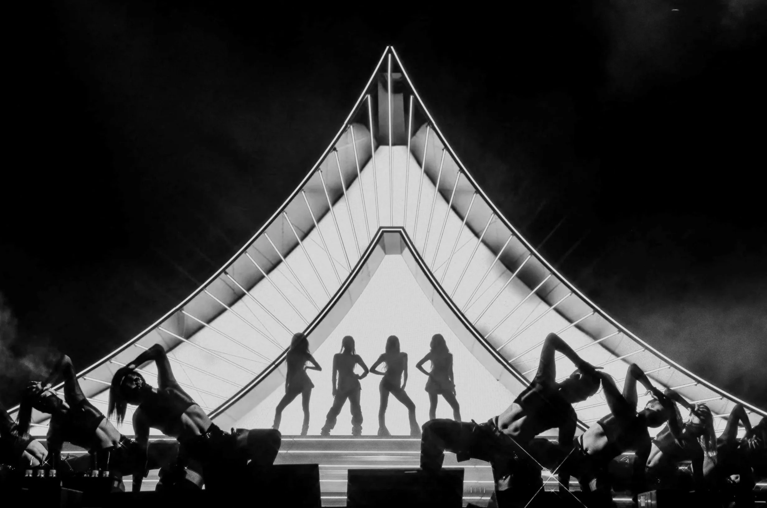 Black and white photo of five women standing on a stage with geometric shapes and lines in the background, seen from below, with silhouettes of people seated and watching.