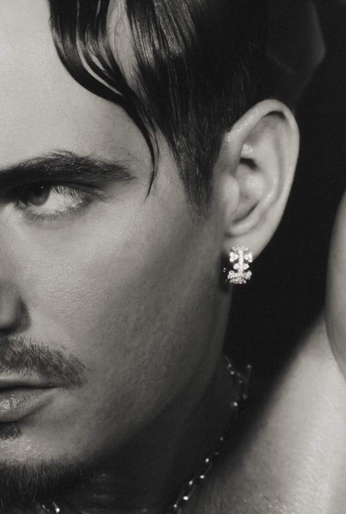Close-up black-and-white photo of a young man with styled dark hair, a mustache, an earring, and a chain necklace, partially showing his face and side profile.
