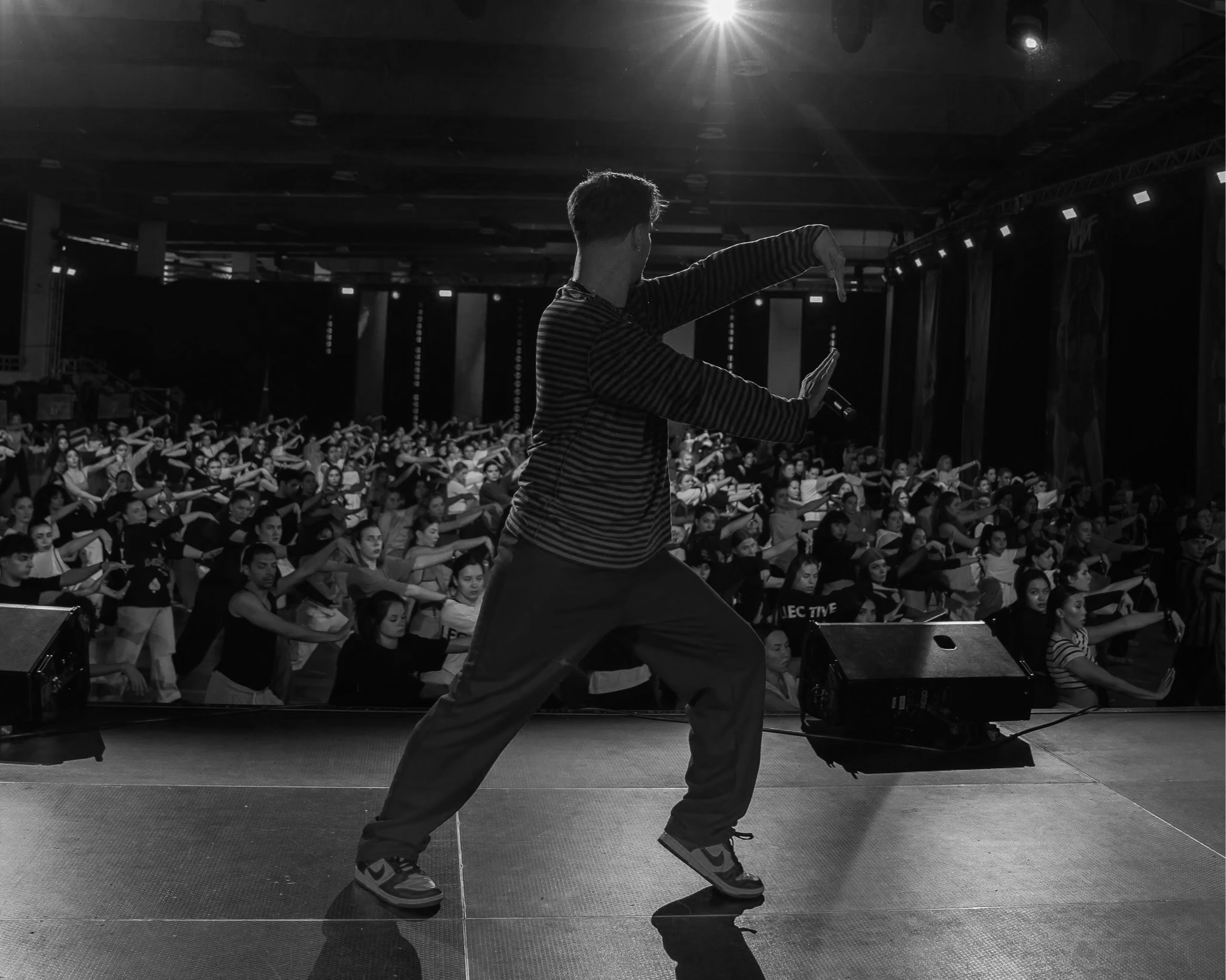 A person dancing in front of an audience on stage in a large hall, with the audience performing synchronized movements, in a black and white photograph.