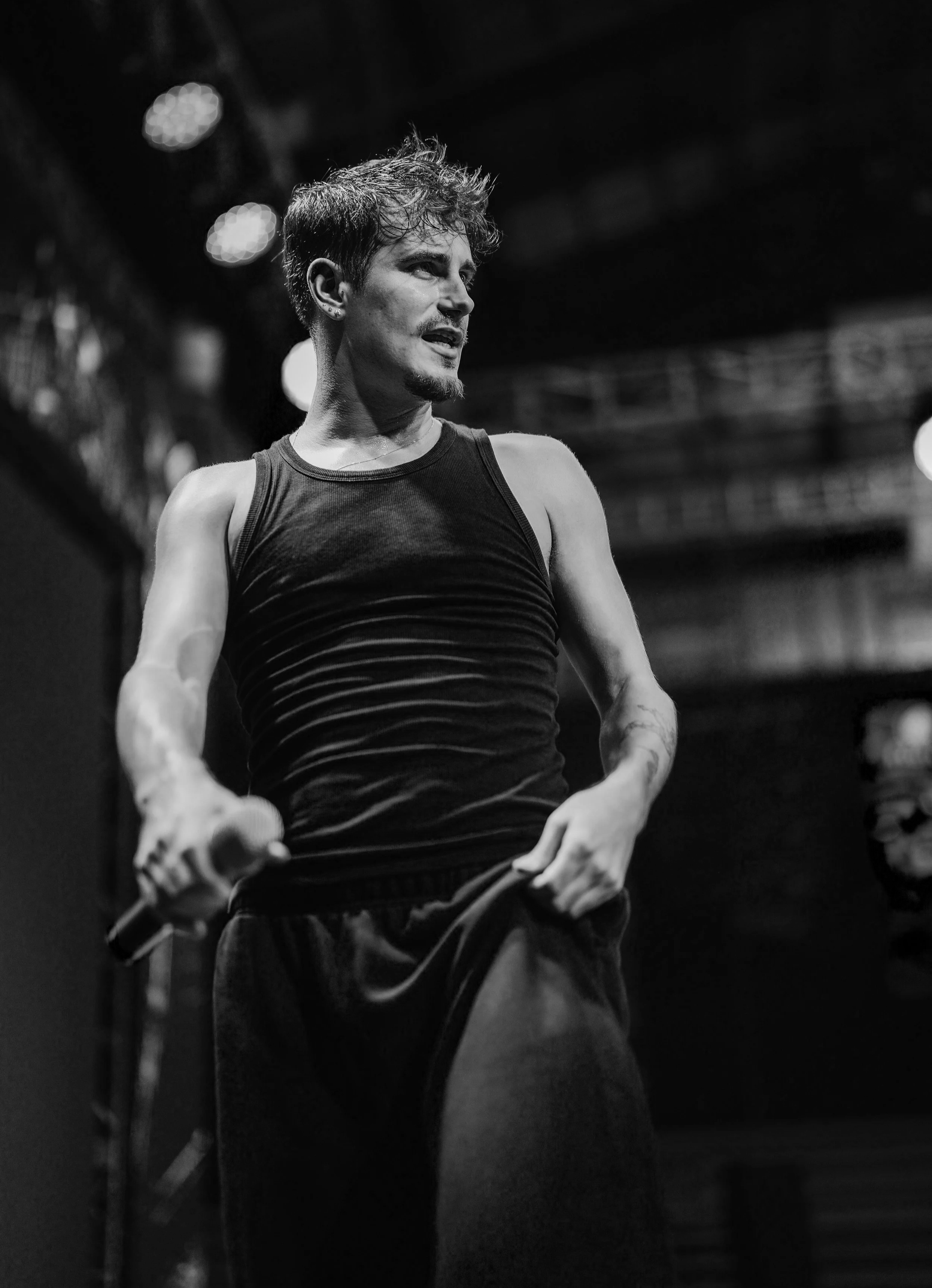 A black and white photo of a young male performer with messy hair, wearing a sleeveless top, holding a microphone, and standing on stage with blurred lights in the background.