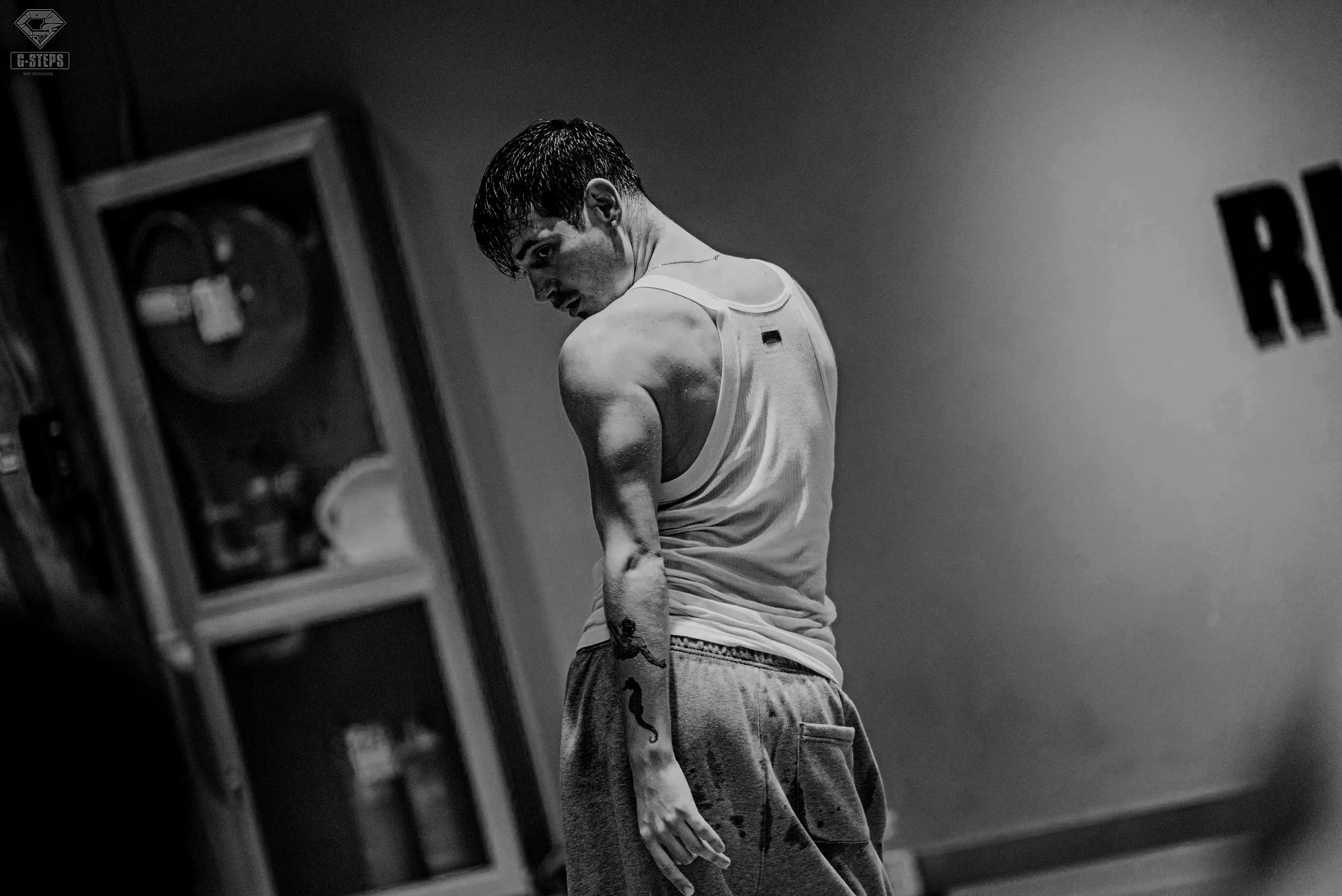 A young man with short dark hair and tattooed arms is standing against a plain wall, looking over his shoulder with a serious expression. He wears a sleeveless shirt and sweatpants. The image is in black and white.