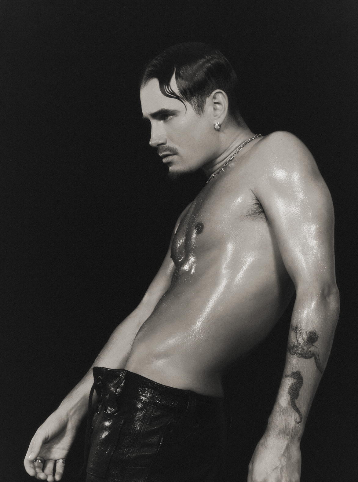 Black and white photograph of a shirtless man with tattoos, wet skin, and dark slicked-back hair, wearing earrings and a chain necklace, standing against a dark background.