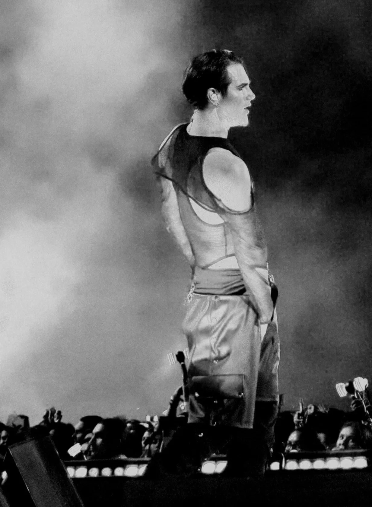 Black and white photo of a woman in a sleeveless top with open shoulders, standing on stage or platform, being watched by an audience.
