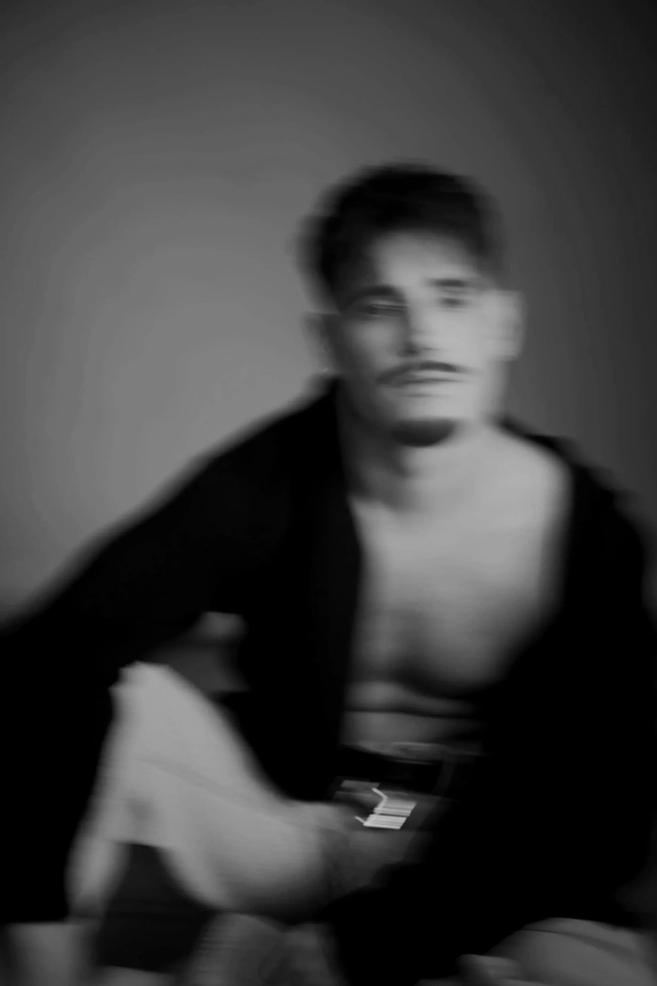 A blurred black-and-white portrait of Kiel Tutin with short dark hair, partially covered by a dark cardigan.
