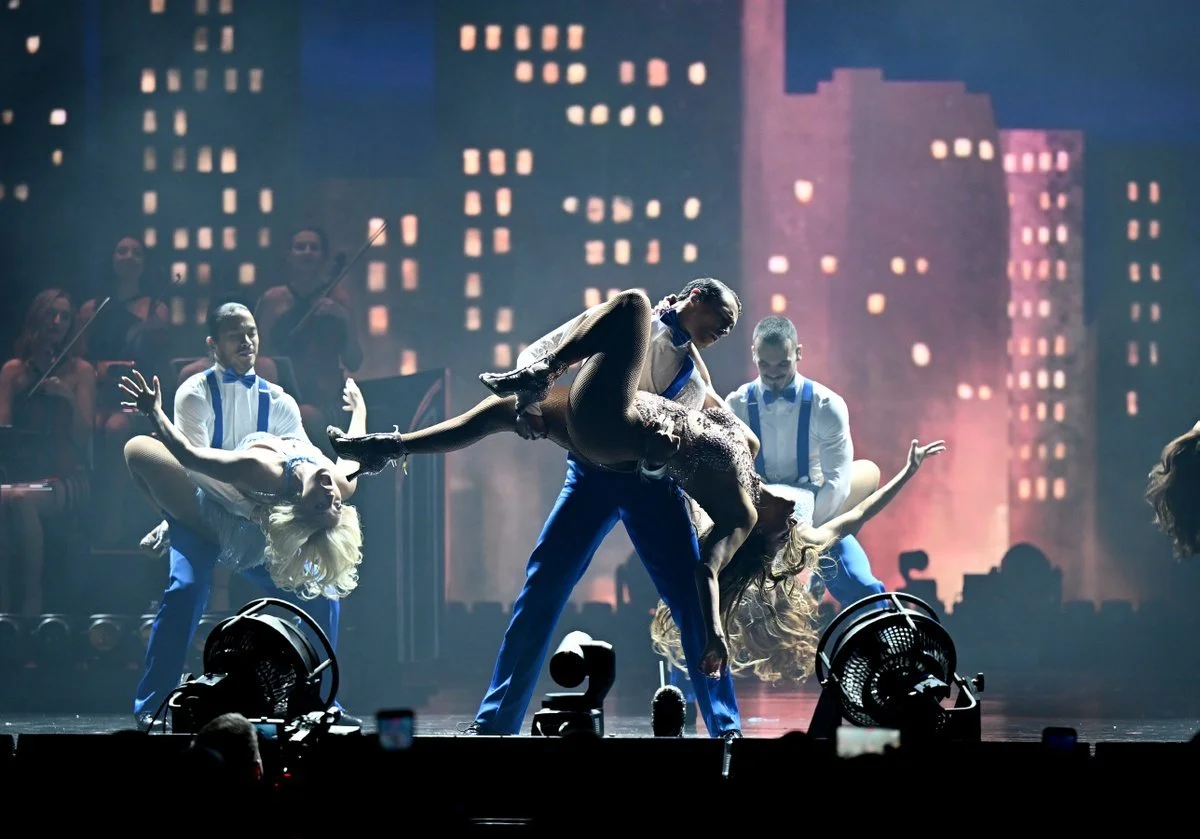 A dance performance featuring four dancers on stage with a city skyline backdrop. The central couple is engaged in a lift with the woman leaning back and the man supporting her. The woman is wearing a sparkling dress, and the others are dressed in coordinated outfits. An orchestra is visible in the background.
