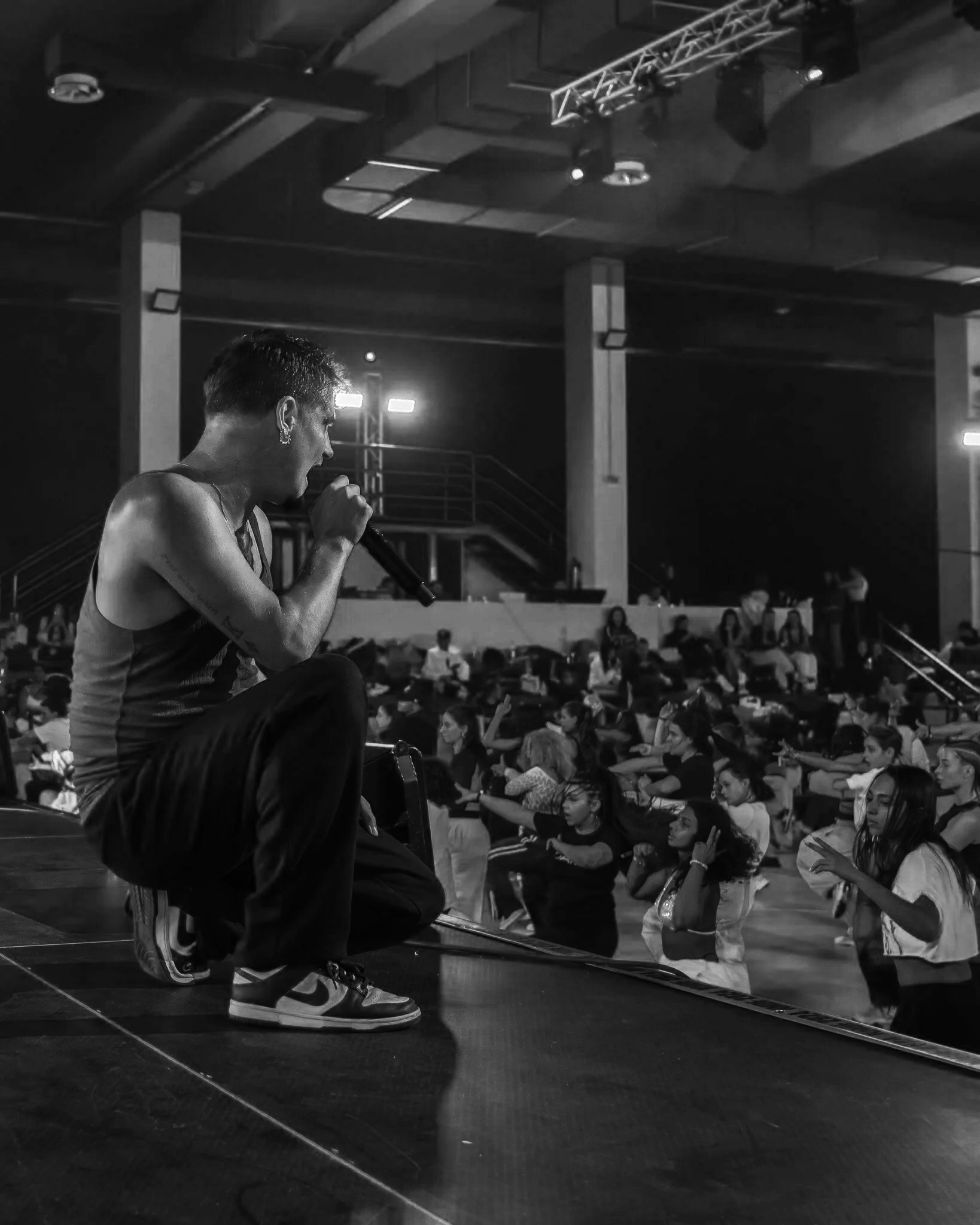 A person crouching on a stage, holding a microphone and speaking to an audience seated on the floor in a dark auditorium or gymnasium.