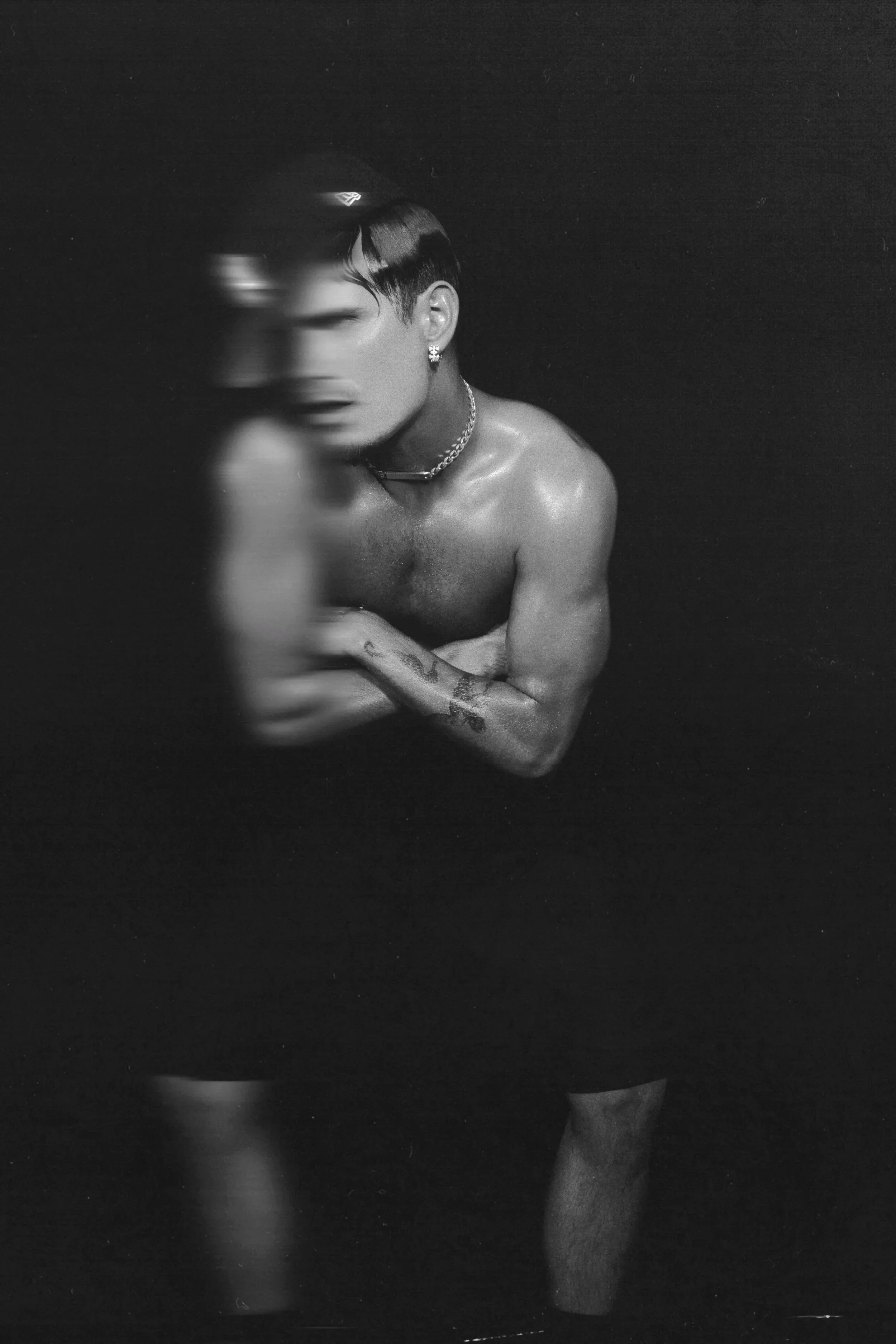 Black and white photo of a shirtless young man with short dark hair, jewelry including a chain necklace and earrings, standing against a dark background, with his face partially blurred and his left arm with tattoos visible.