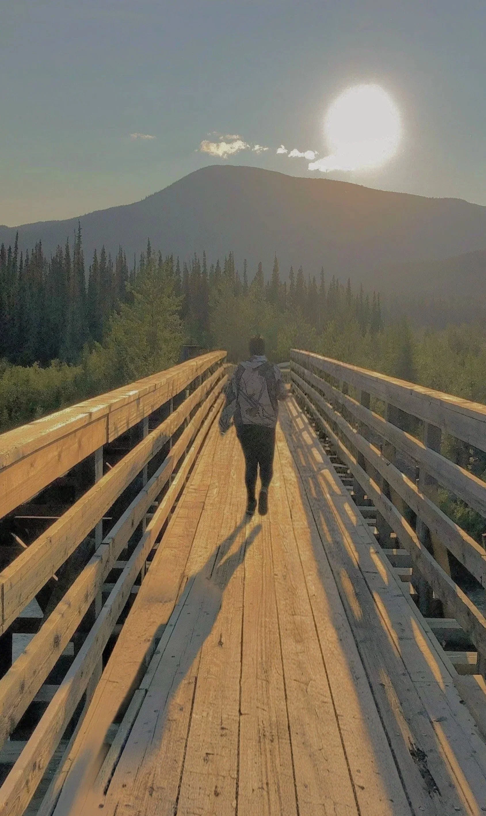Person walking on a wooden bridge towards a mountain with the sun shining brightly in the sky.