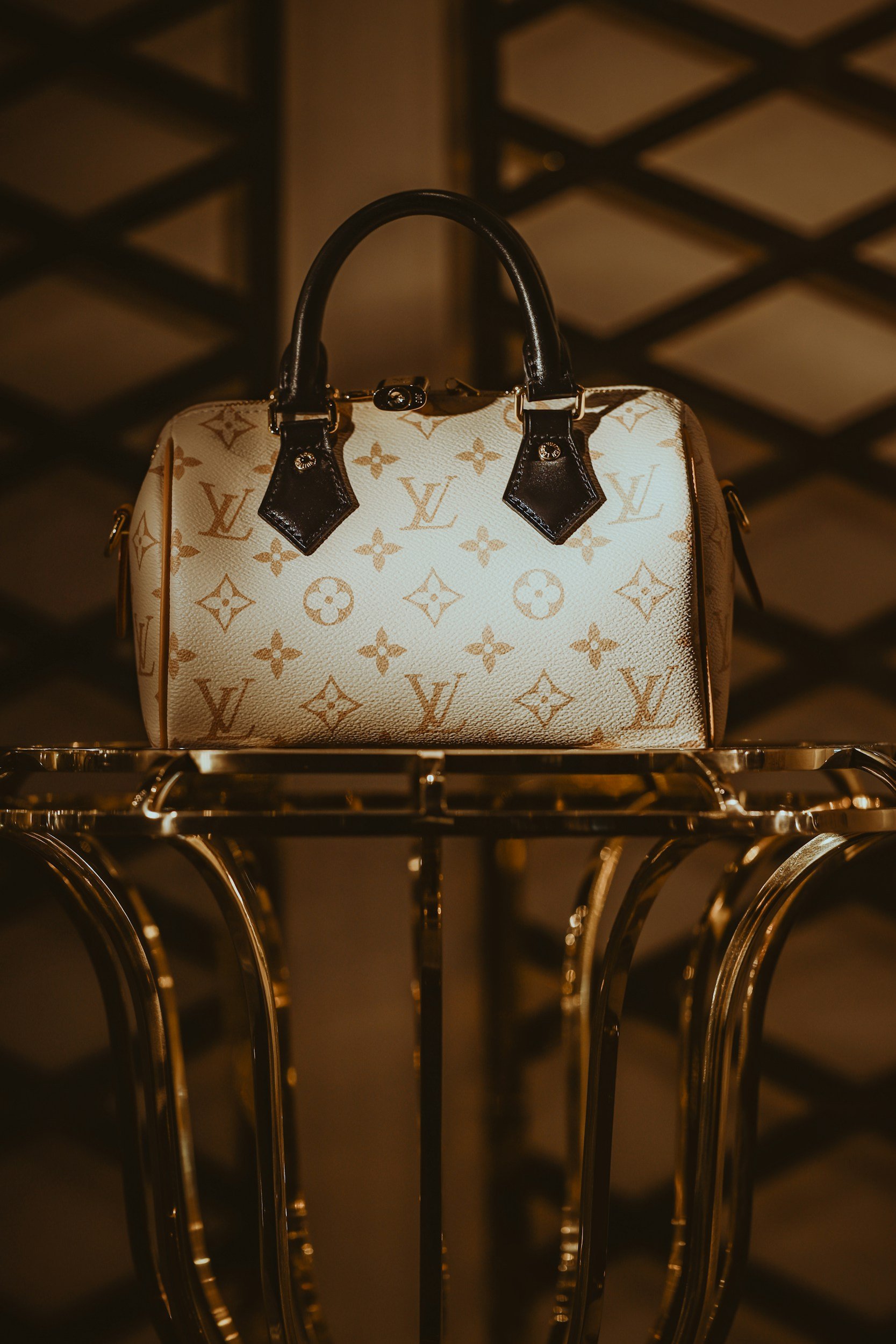 A Louis Vuitton handbag with the brand's monogram pattern, placed on a shiny metallic surface with a dark, blurred background featuring a lattice design.