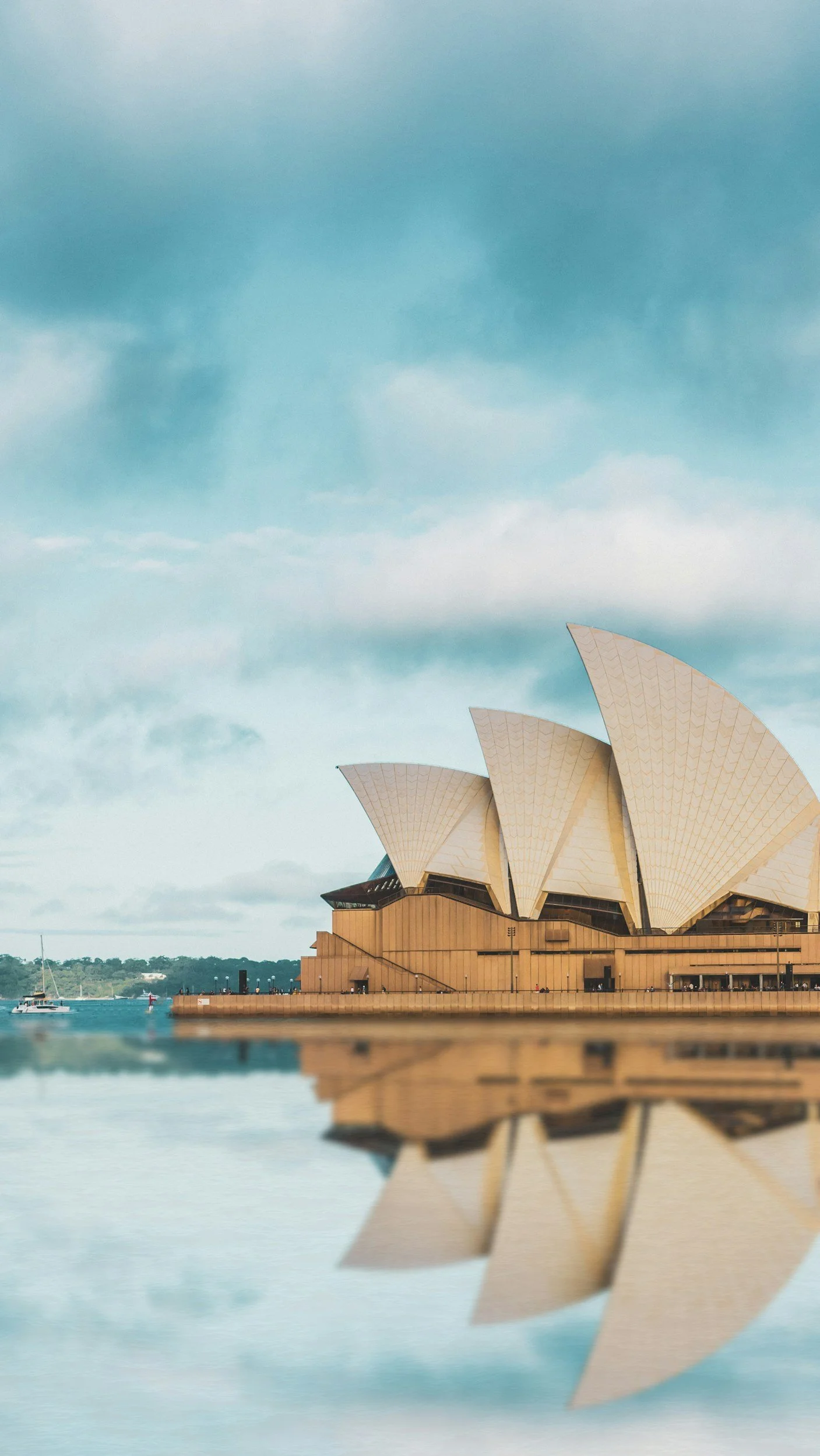 Sydney Opera House with its distinctive sail-like architecture, situated on the water with a boat sailing nearby and its reflection visible in the calm water.
