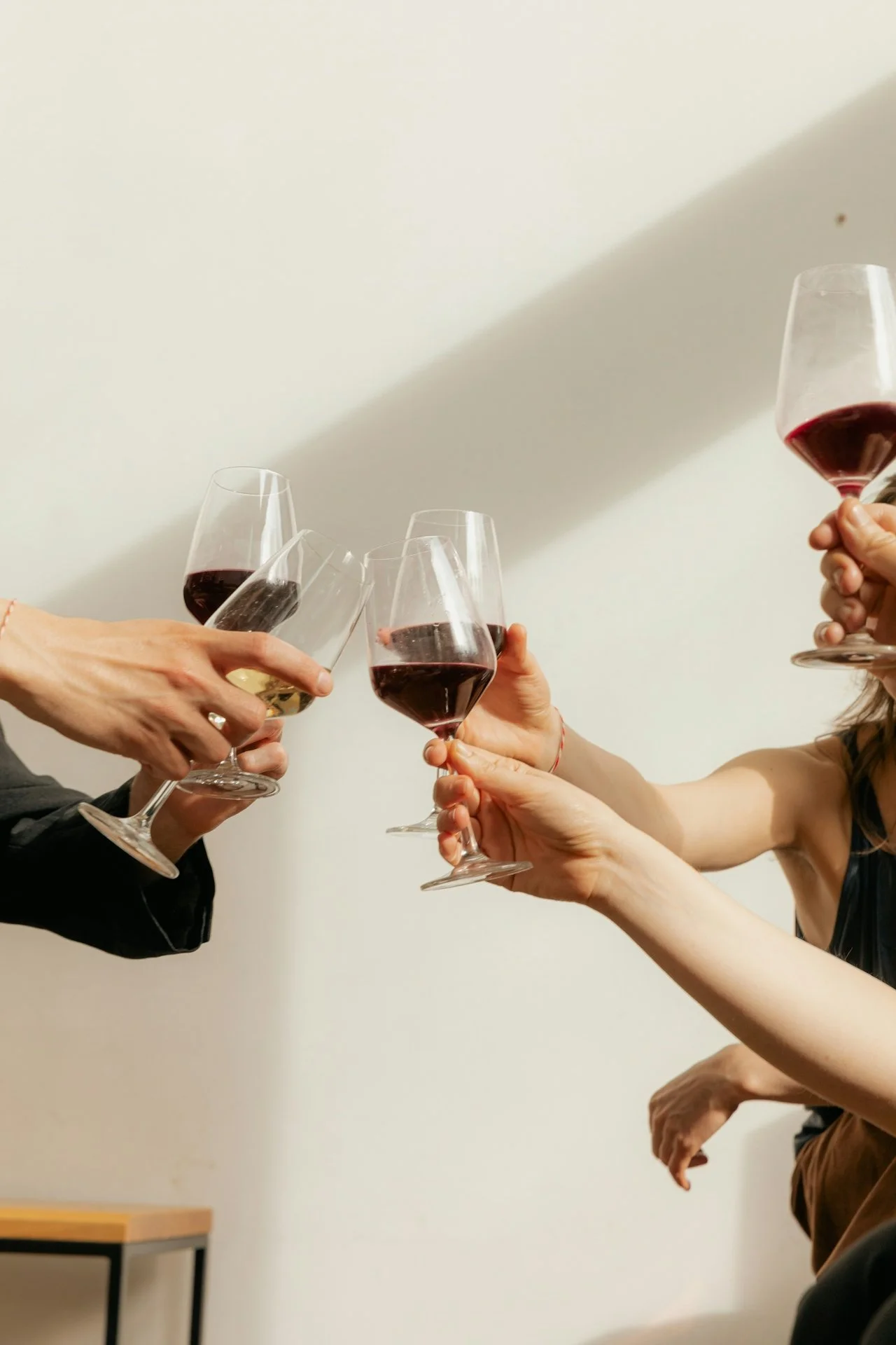 People raising glasses of red and white wine in a toast against a plain wall.