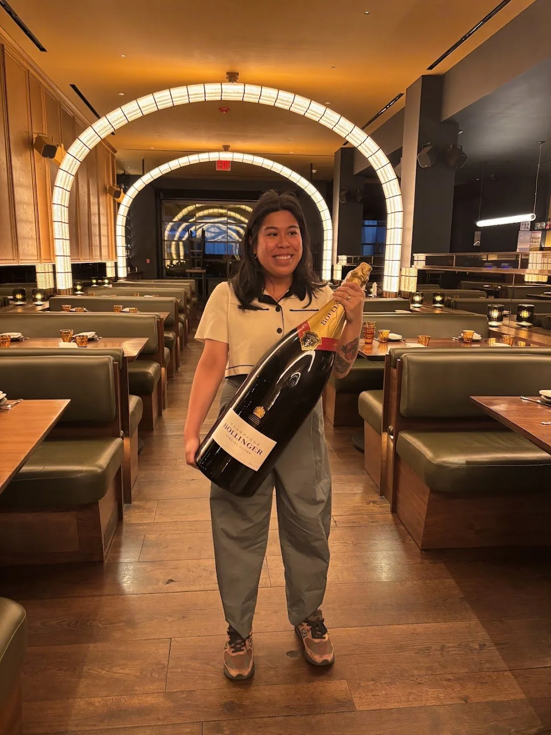 A woman smiling and holding a large bottle of Bollinger champagne inside a restaurant with wooden tables, green booths, and modern lighting fixtures.