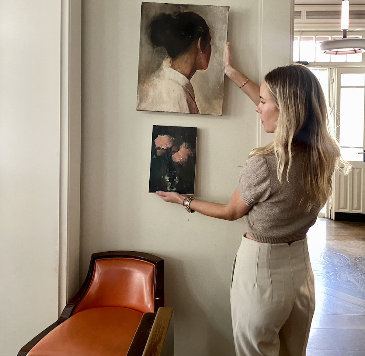 A woman with long wavy blonde hair, wearing a brown knit top and beige pants, is hanging and holding up two paintings on a light-colored wall in a room. One painting features a woman's profile with her hair in a bun, and the other is a floral still life of pink flowers.